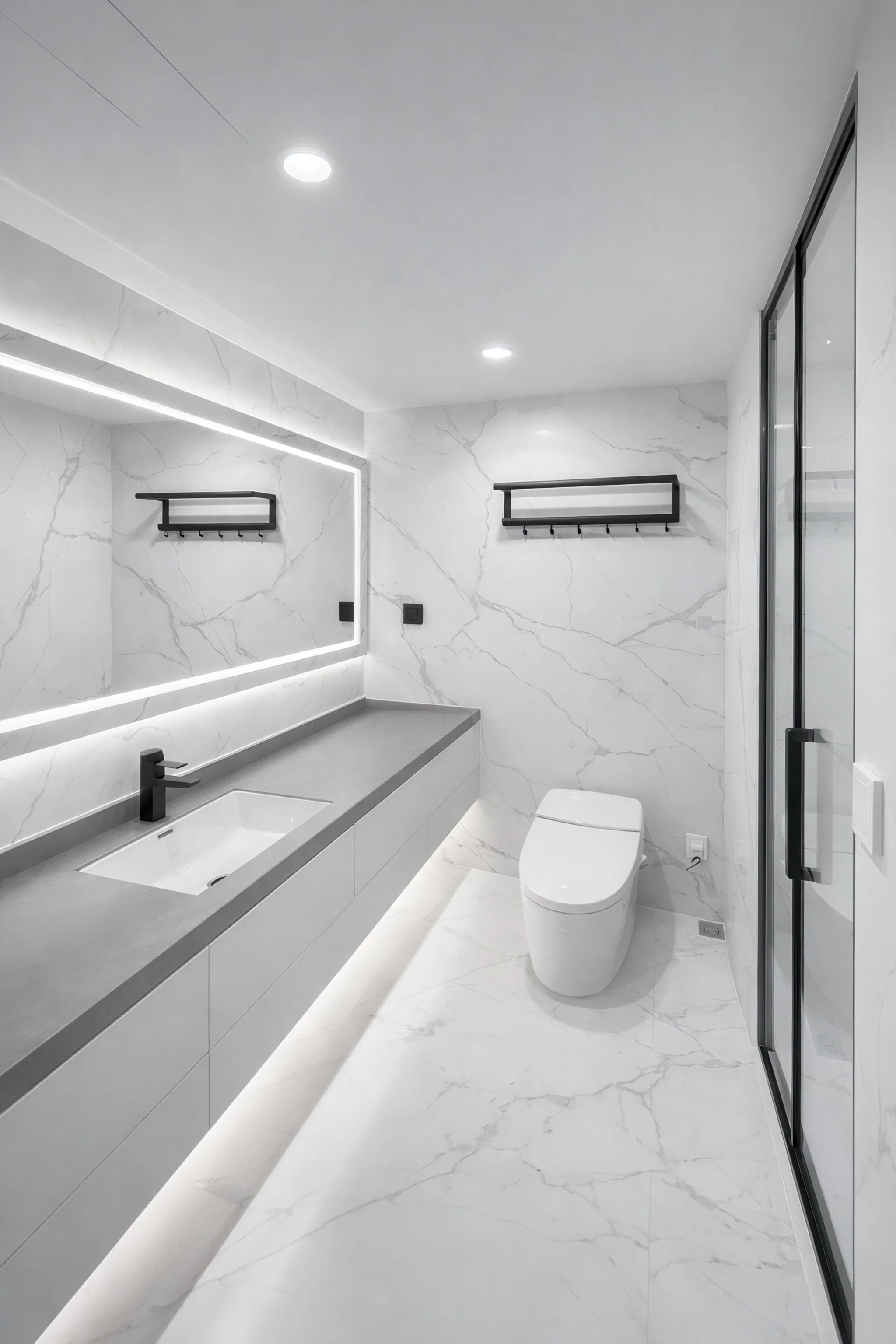 Modern bathroom with white marble walls and floor, black fixtures, a large mirror with LED lighting, a gray countertop with an integrated sink, black wall-mounted shelves with hooks, a toilet, and a glass shower door.