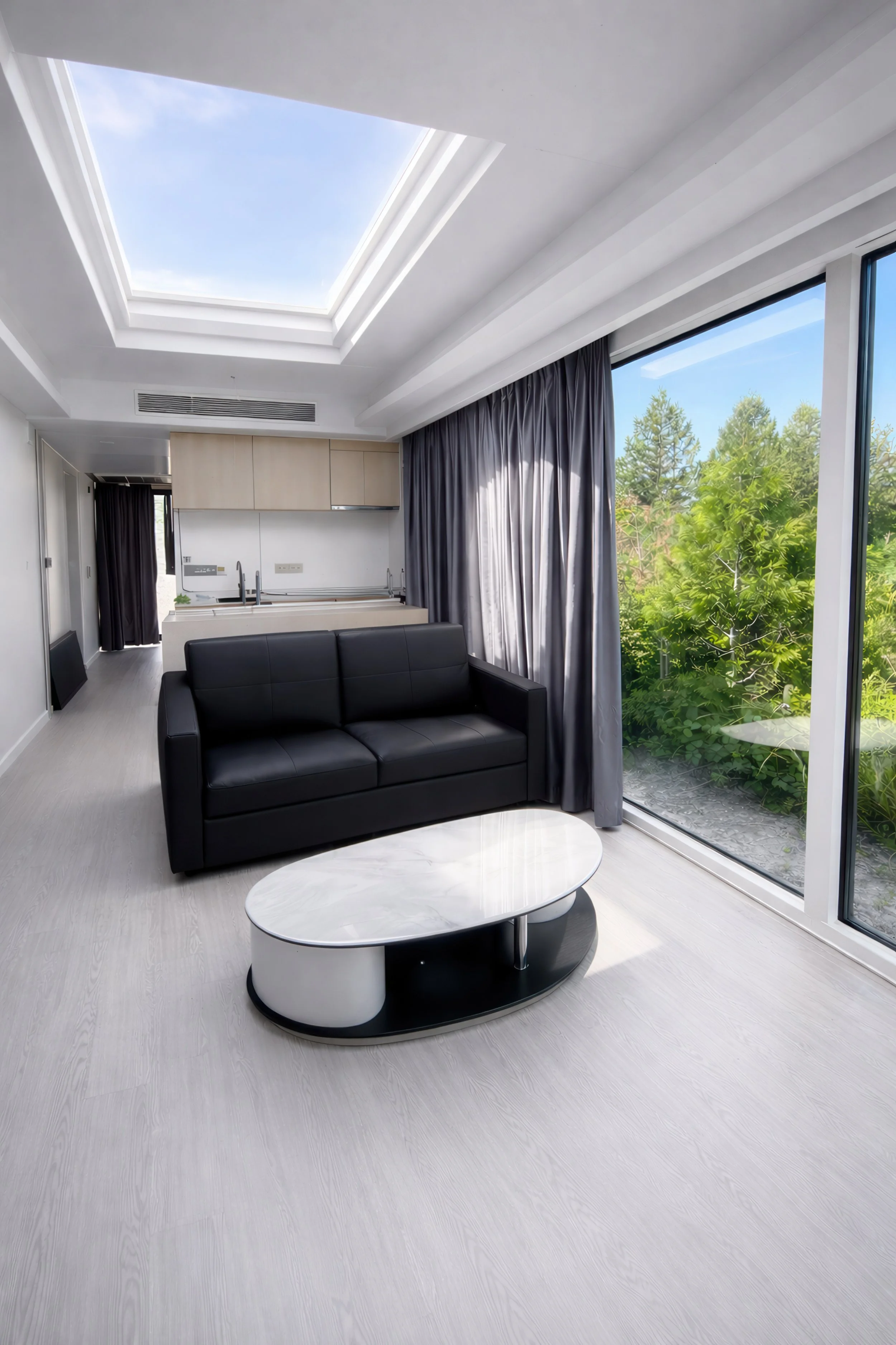 Living room with a black sofa, a white marble oval coffee table, large windows with dark curtains, a kitchen area with wooden cabinets, and a skylight with a view of blue sky and trees outside.