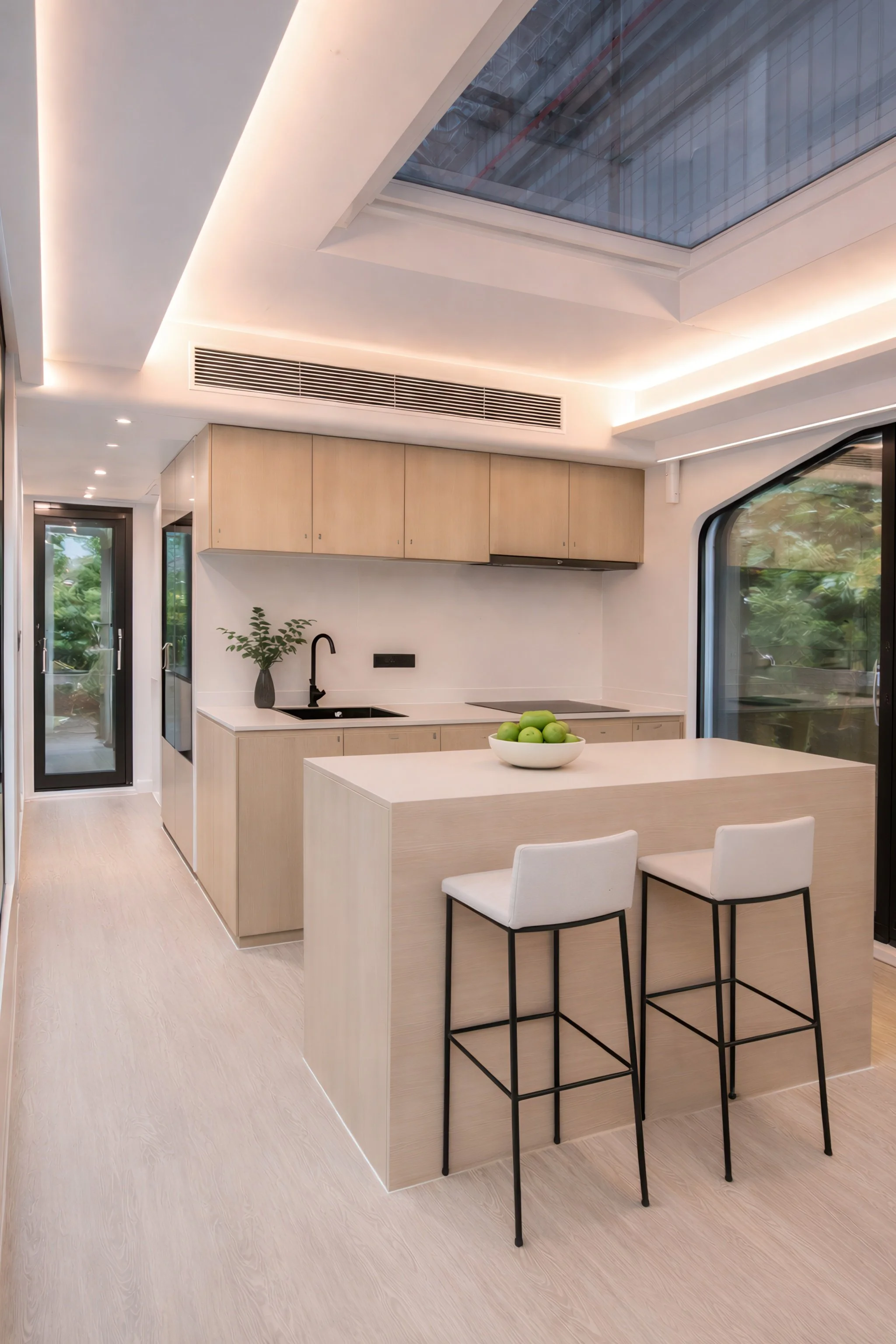 Modern kitchen with light wood cabinetry, white countertops, a black sink, two white bar stools, a bowl of green apples, large windows, and a skylight.