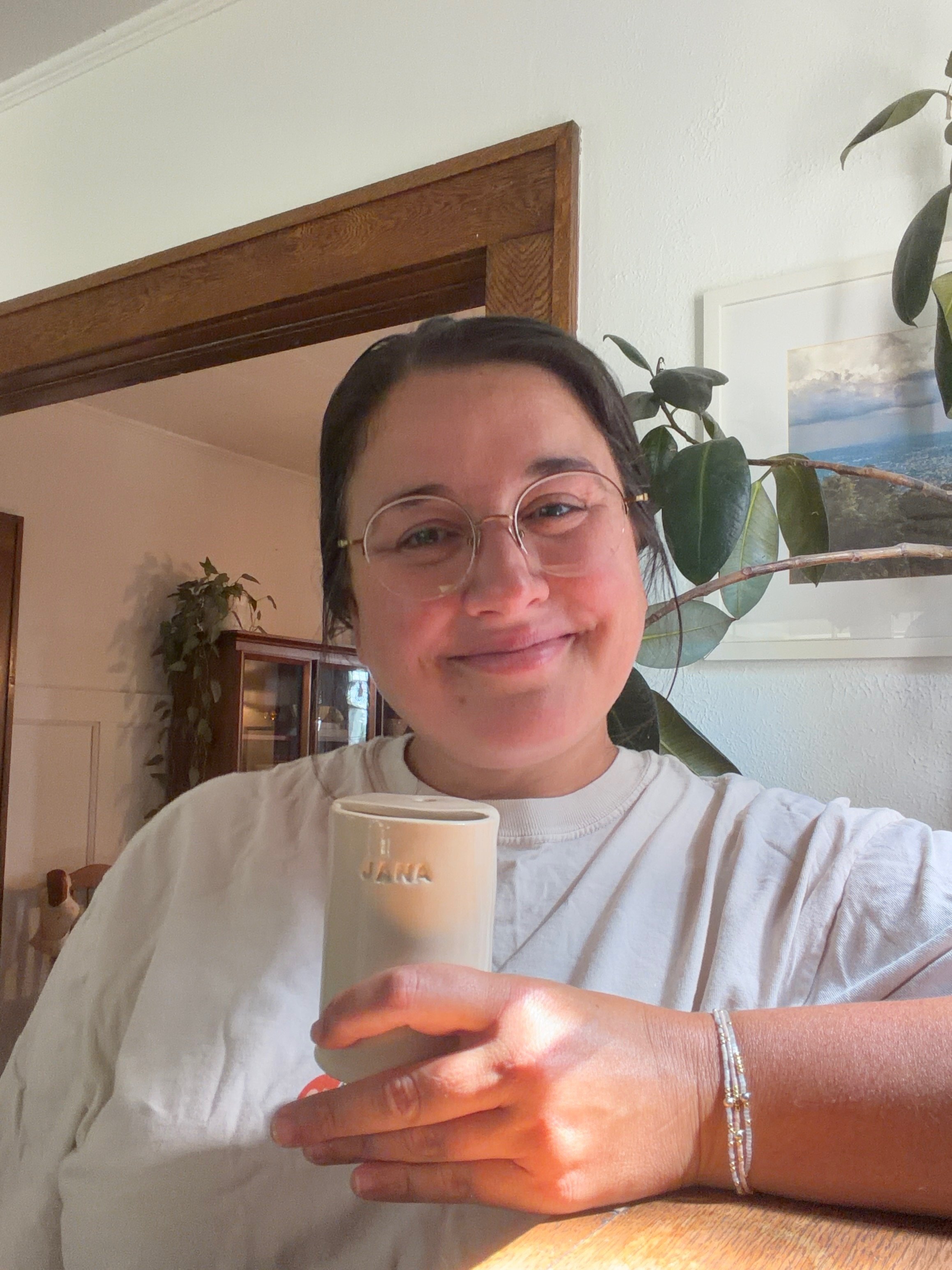 A woman smiling, wearing glasses and a white shirt, holding a beige mug in a cozy home setting with plants and wall art.