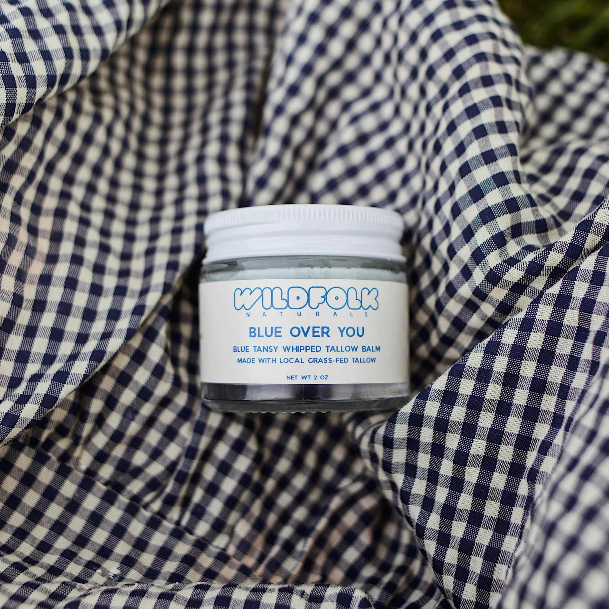 A person wearing blue and white checkered shirt, sitting outdoors, holding a white container labeled 'Wilfreds Naturals Blue Over You' with blue-tansy whipped tallow balm.