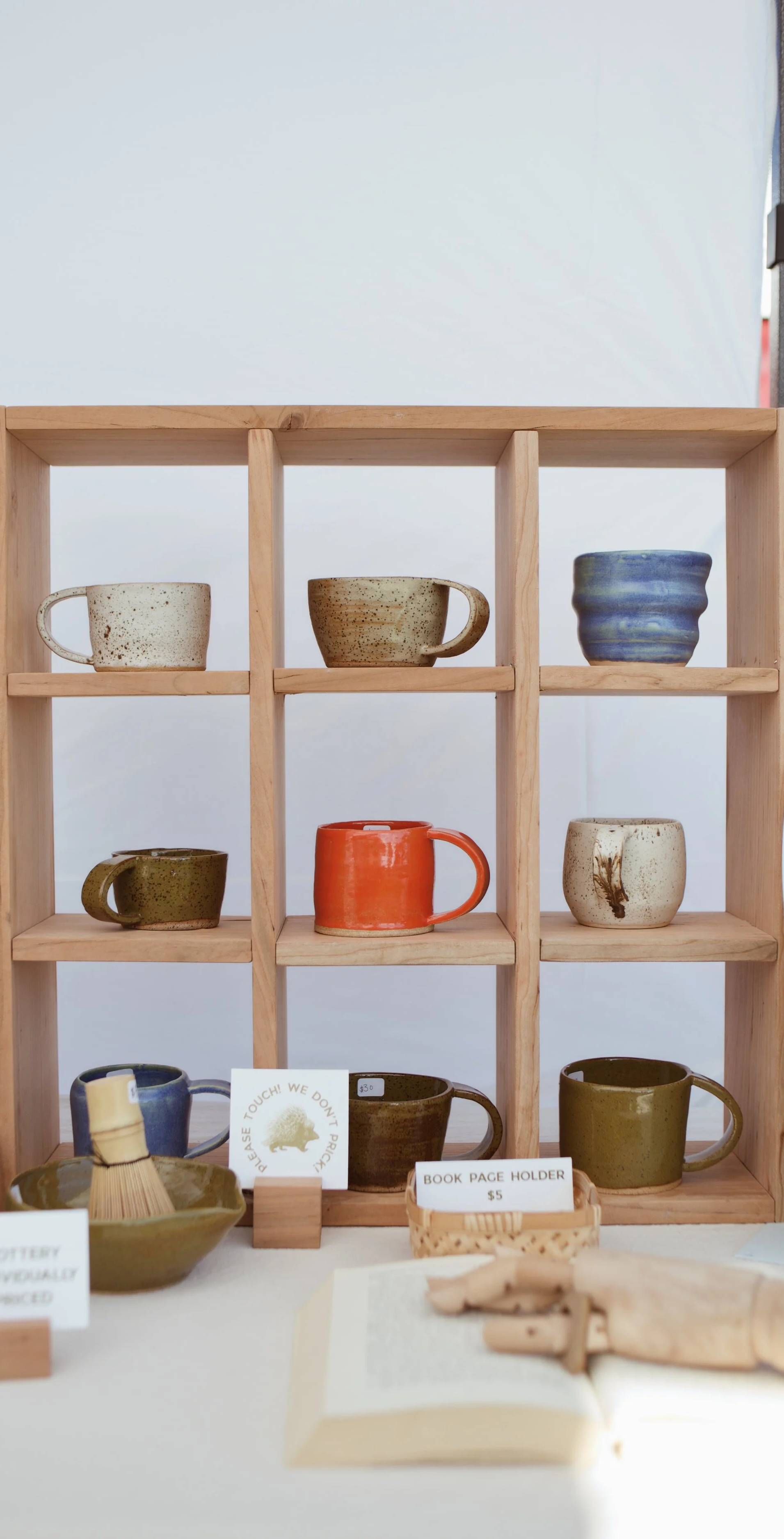 Display of various pottery cups and mugs arranged on a wooden shelf and table, with price tags and small signs.