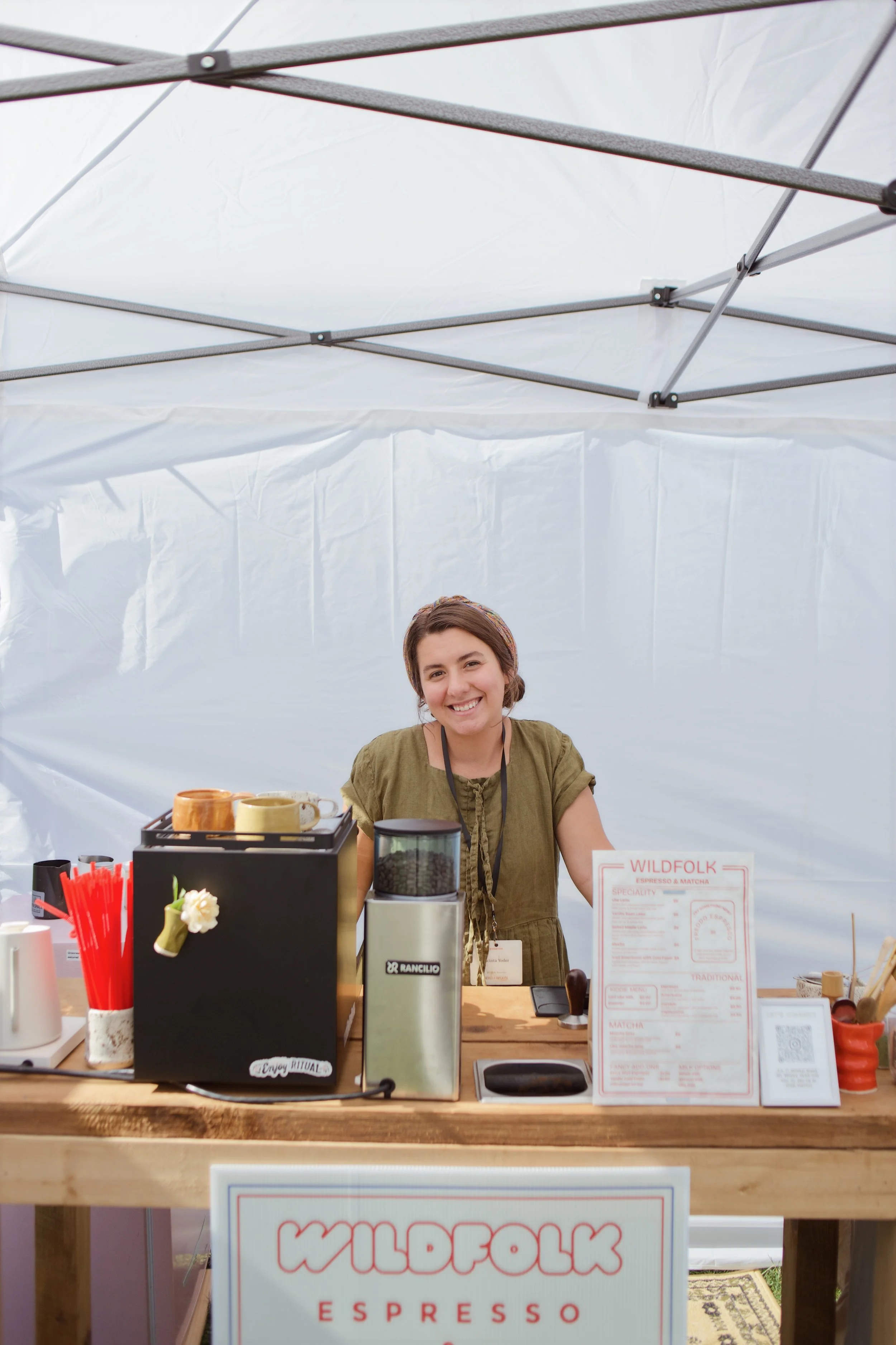 Smiling woman standing behind a coffee stand at an outdoor event, with various coffee-making equipment and a menu on display.