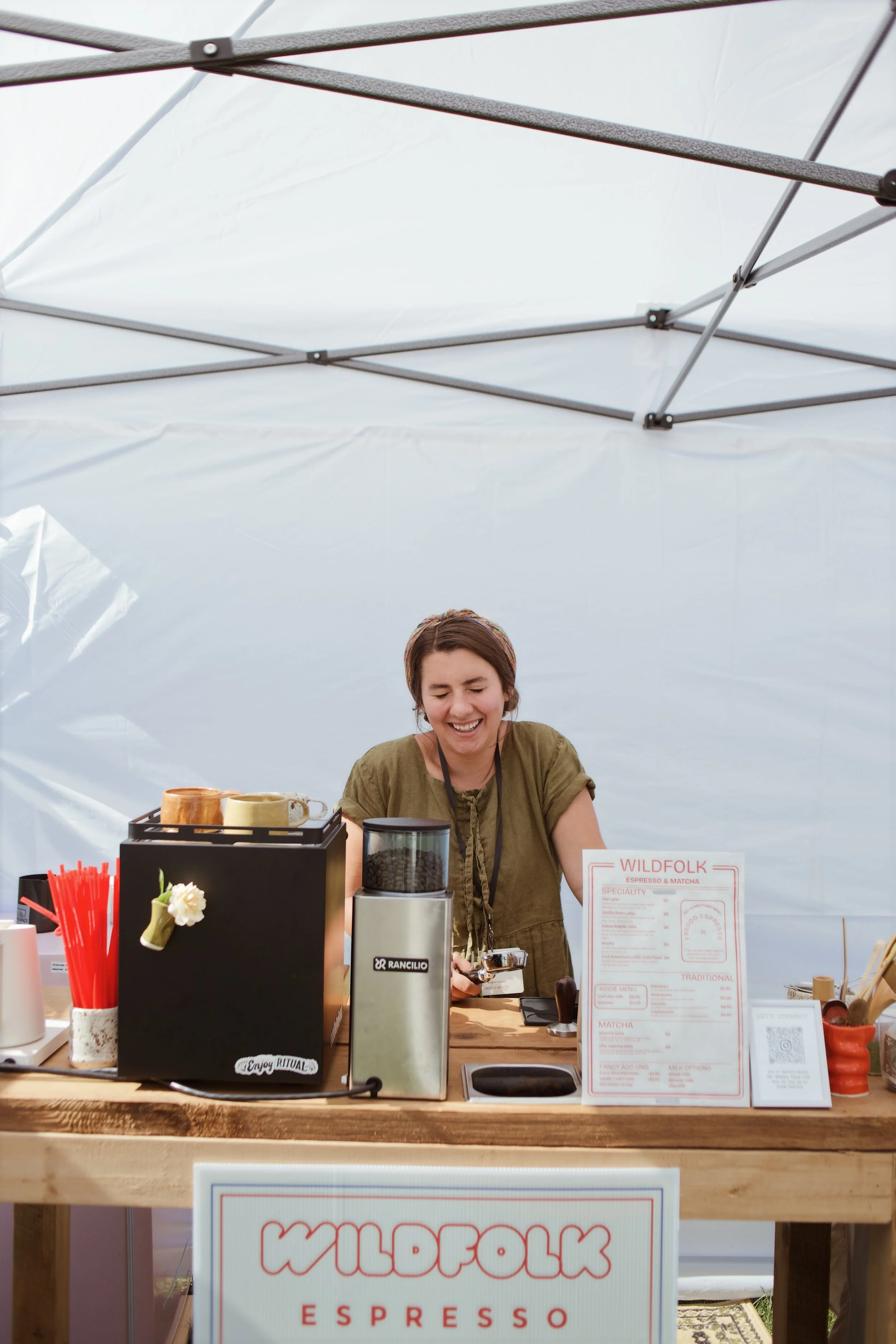 A smiling woman behind a wooden counter at a coffee stand, with a large sign that says "Wildfolk Espresso" and a menu displayed on the side, under a white canopy.