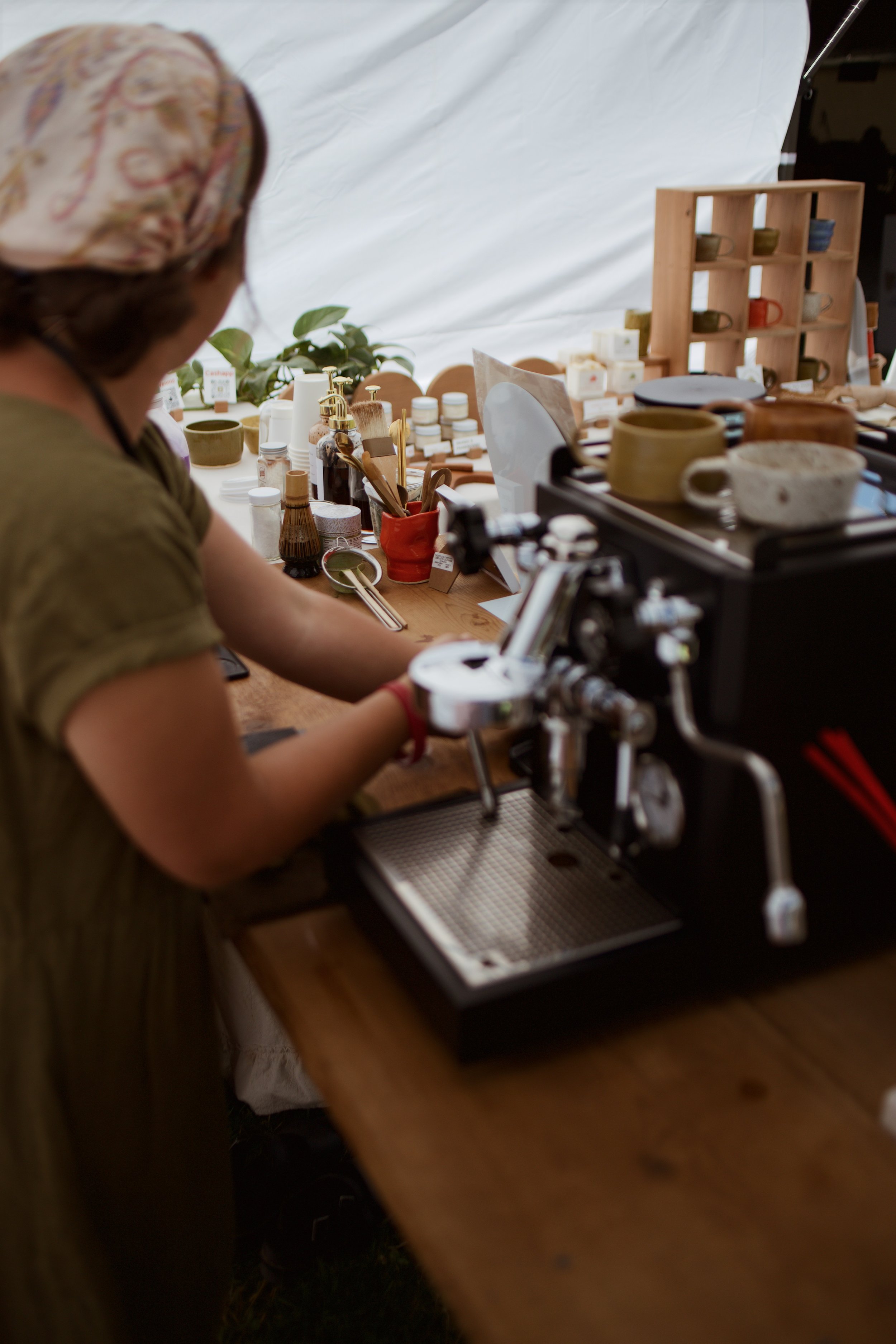 A person wearing a headscarf working at a wooden table with pottery and soap-making supplies, such as brushes, bottles, and soap bars, in a craft or art studio.