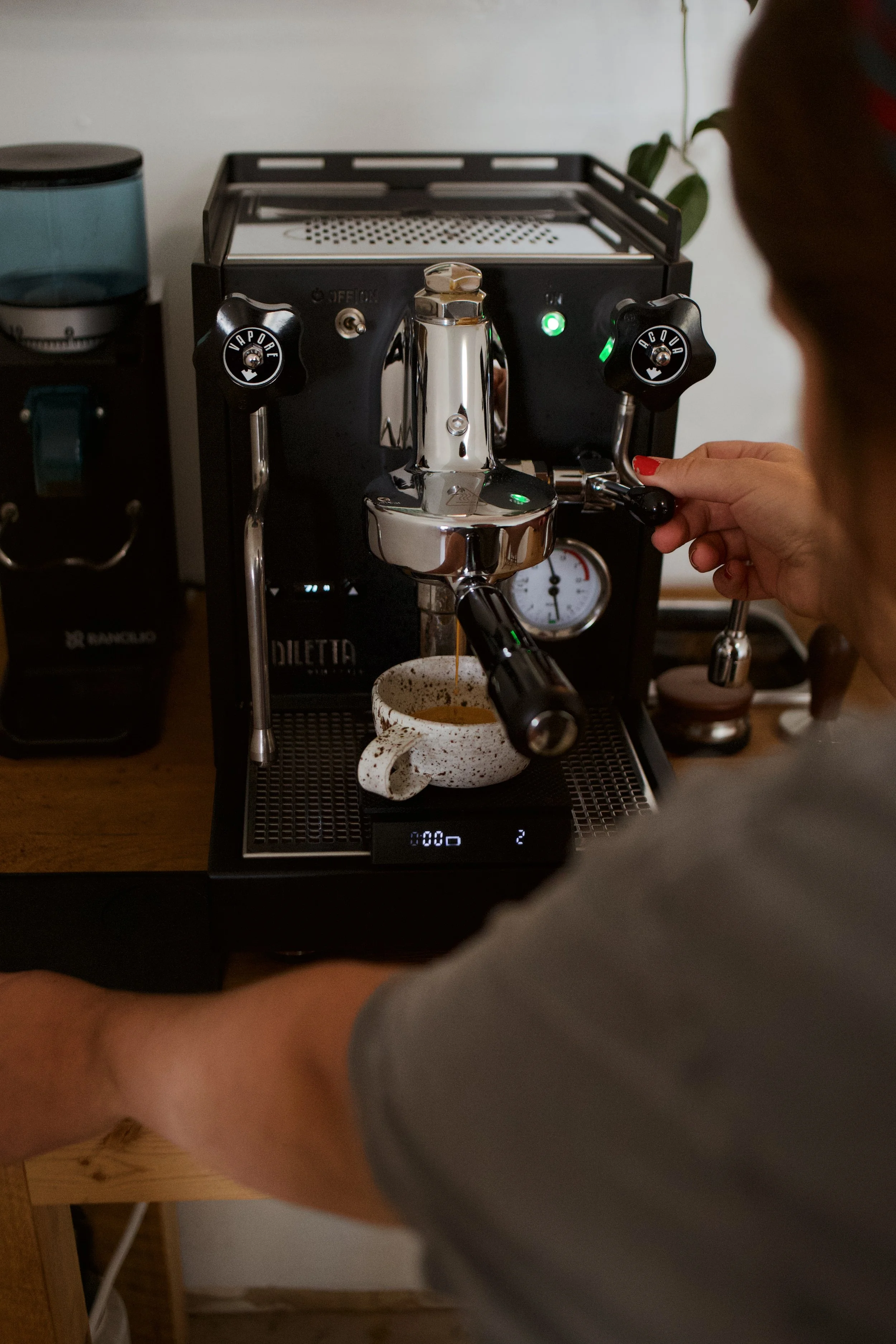 A person is operating a coffee machine, pouring hot water into a portafilter, with a coffee mug positioned underneath. The scene appears to be in a kitchen or coffee brewing area.