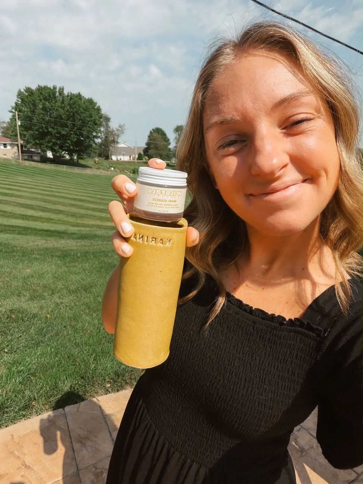 A woman with blonde hair smiling outdoors, holding a yellow KABIN bottle and a small jar of Mamacare skincare cream. She is standing on a patio with a grassy field and houses in the background, under a partly cloudy sky.