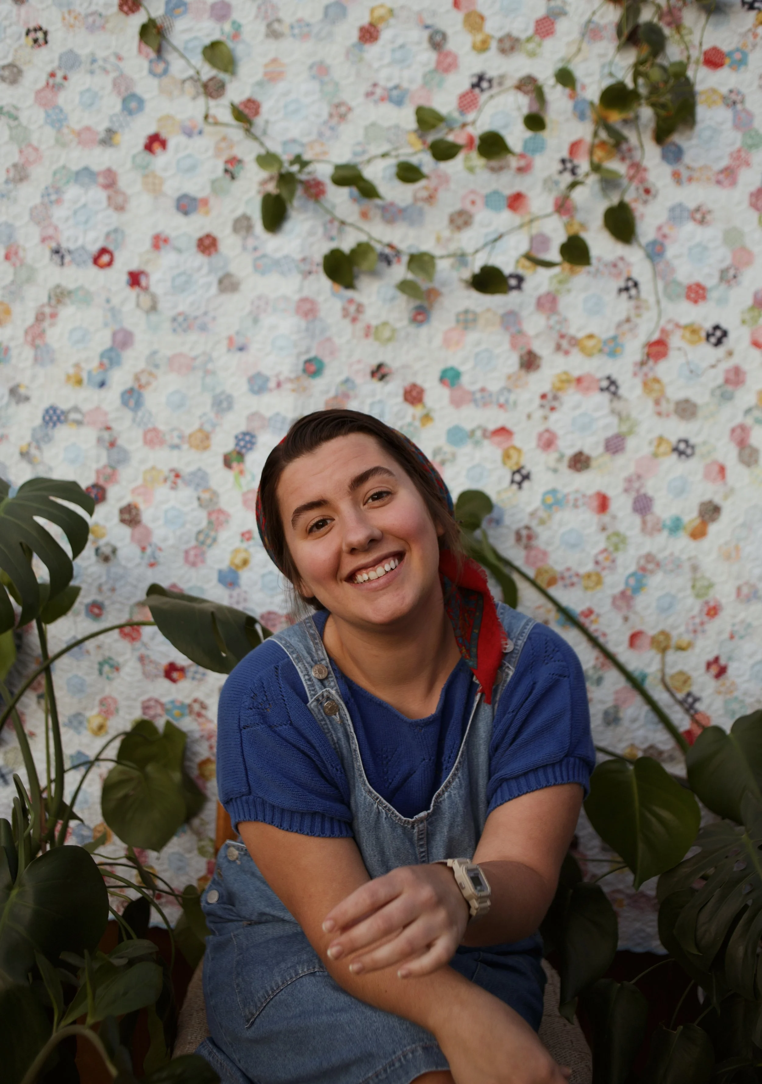 A smiling young woman with short dark hair, wearing a blue shirt and denim overalls, sitting among green plants with large leaves, in front of a colorful quilted background.
