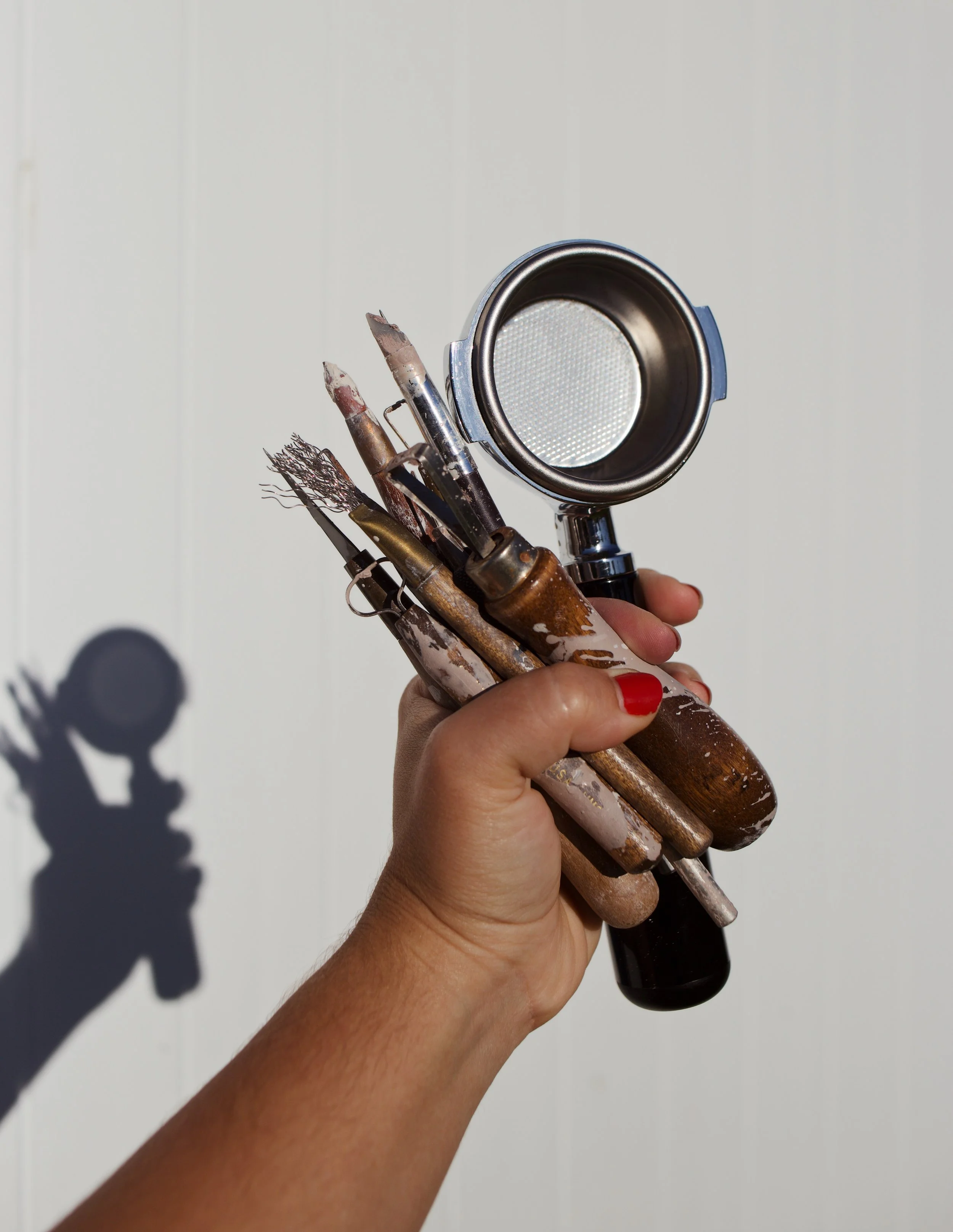 A hand holding several paintbrushes and a paint spray gun against a plain white wall, with the shadow of the person visible on the wall.