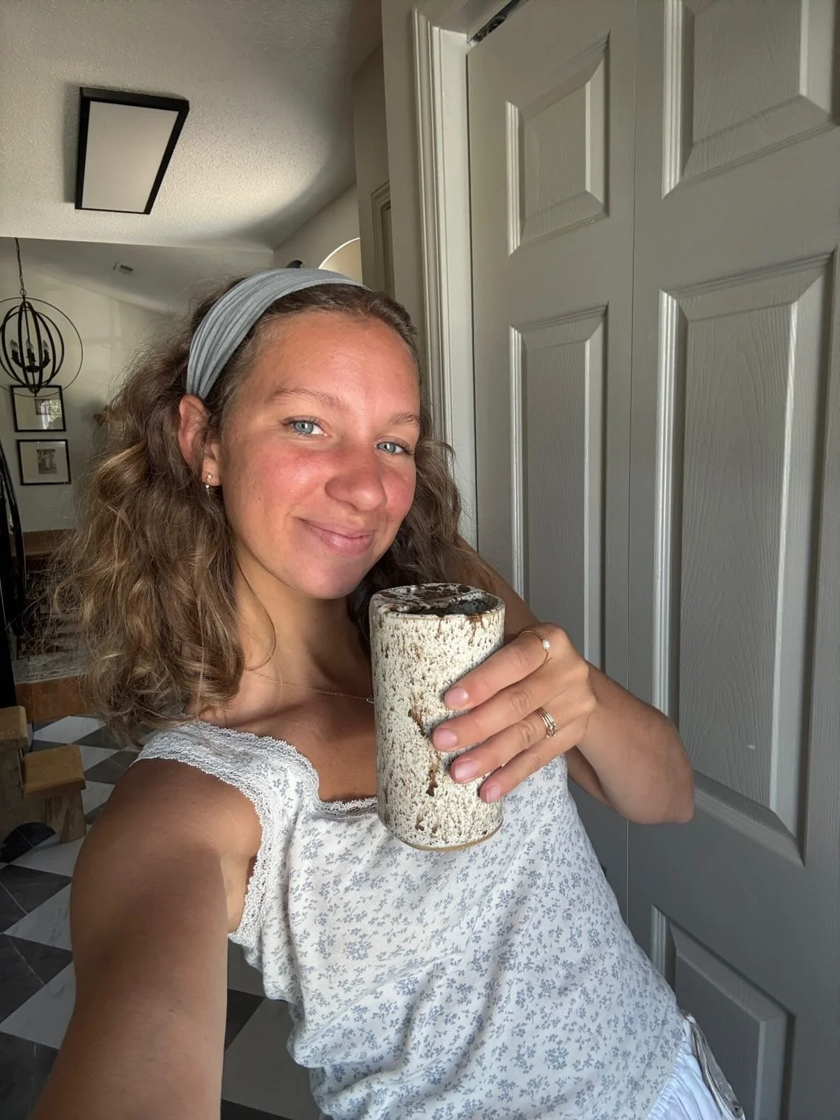 A woman with curly hair wearing a light gray headband and a white sleeveless top, smiling while holding a ceramic mug with a rustic, speckled design, standing inside a room with a white door and modern decor.