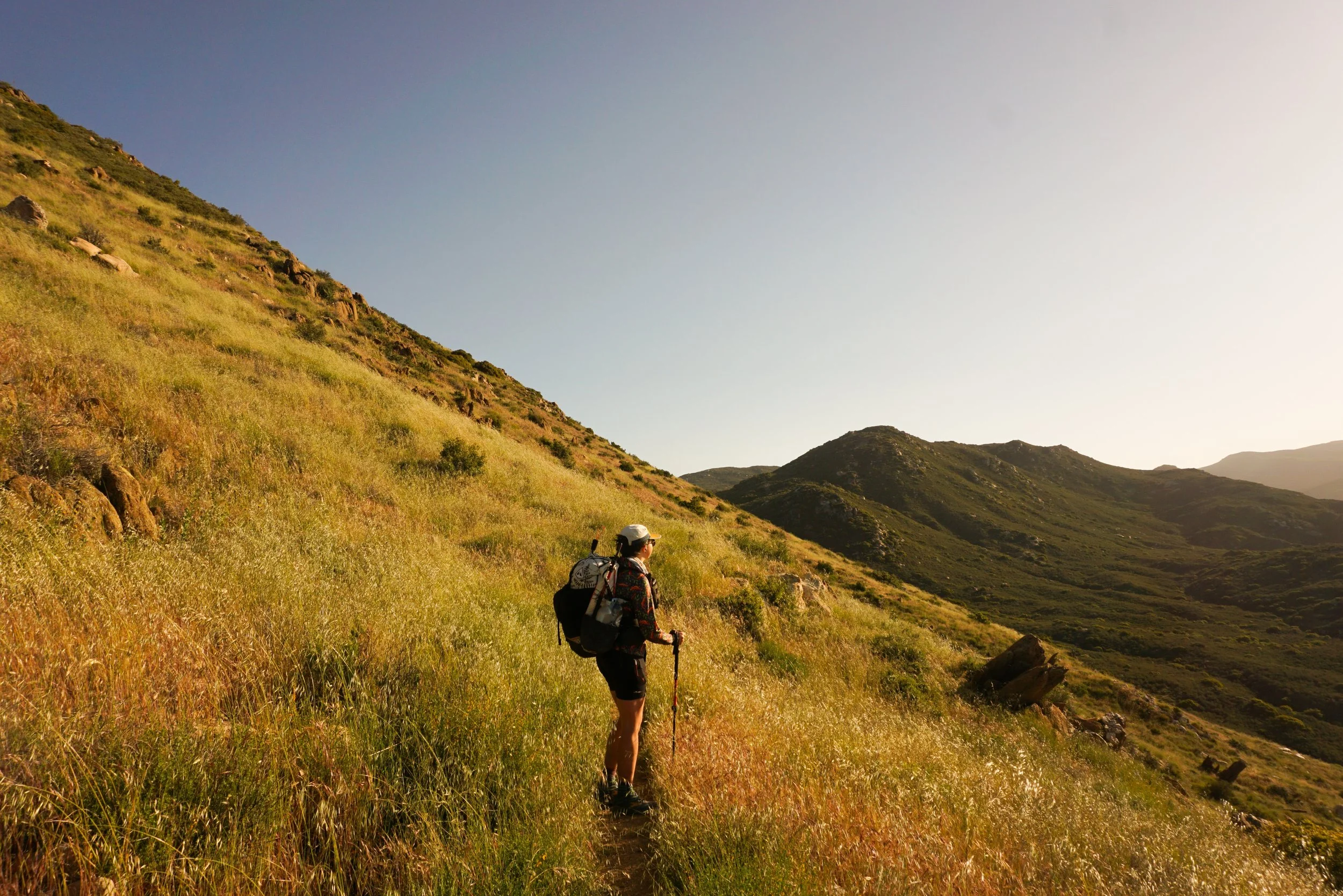 Hiker walking on a trail through a grassy, mountainous landscape during sunset.