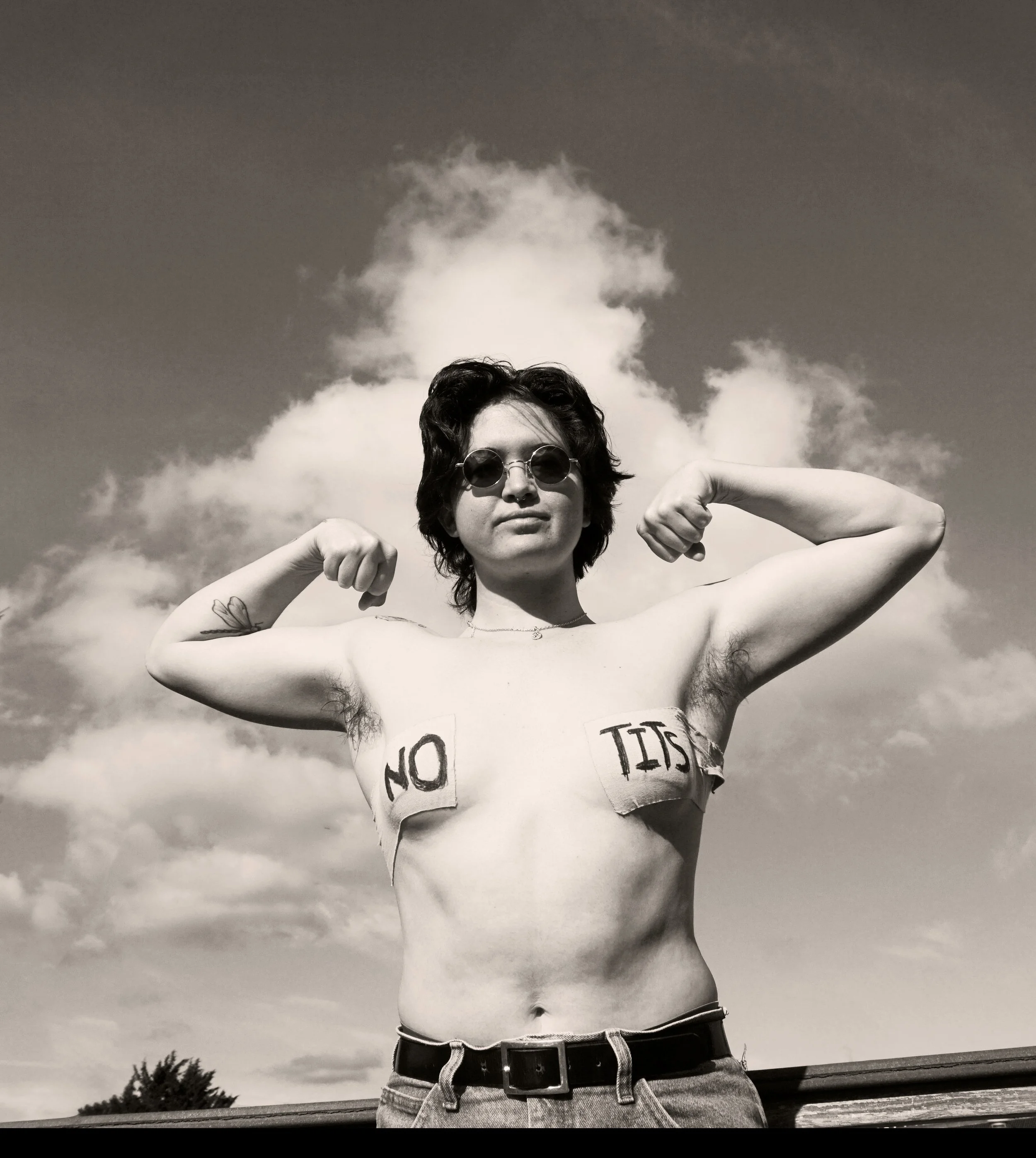 Black and white photo of a person with short hair, sunglasses, and tattoos, flexing their biceps with the words 'NO' on one breast and 'TISS' on the other.