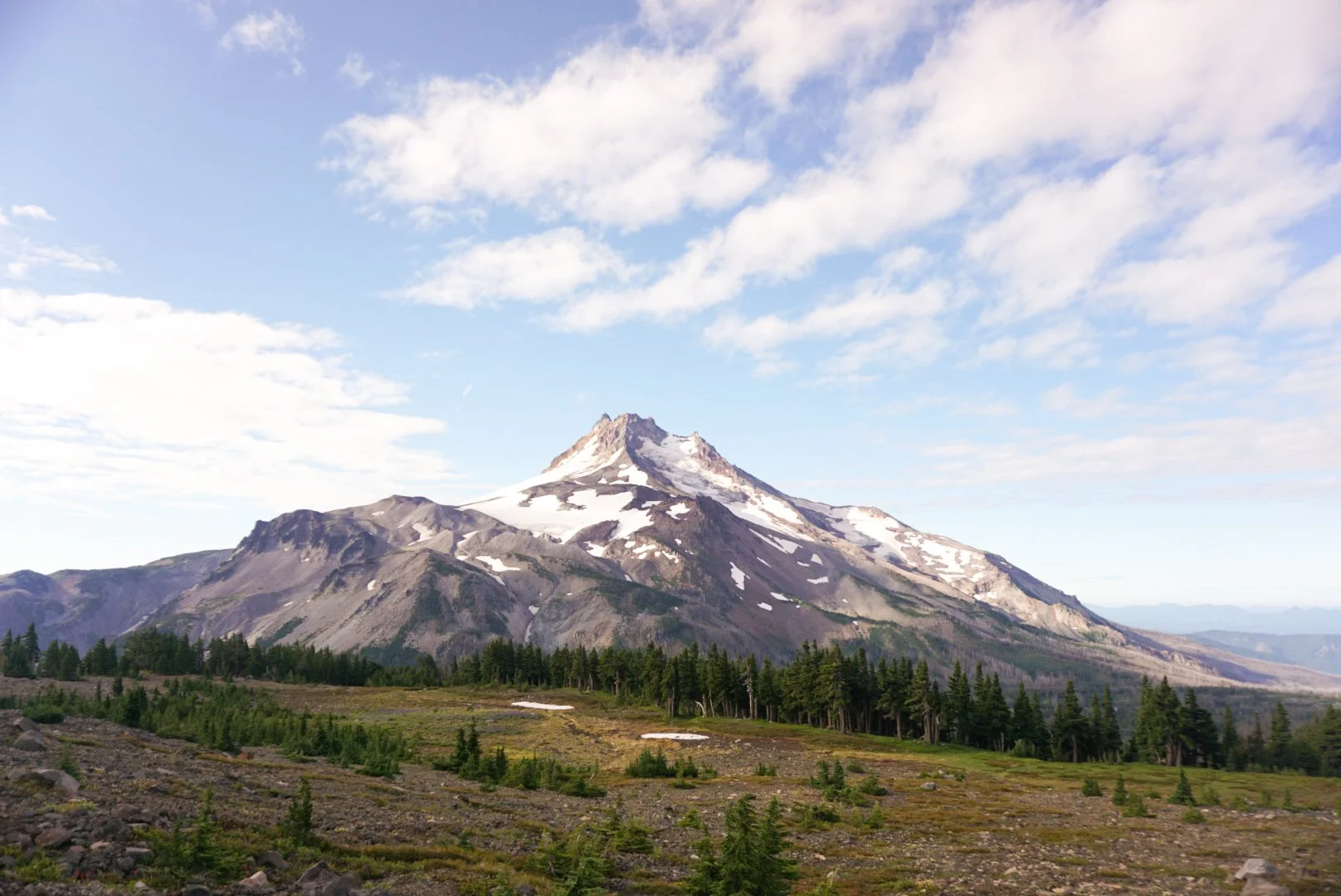 A snow-capped mountain surrounded by green forests with a cloudy blue sky.