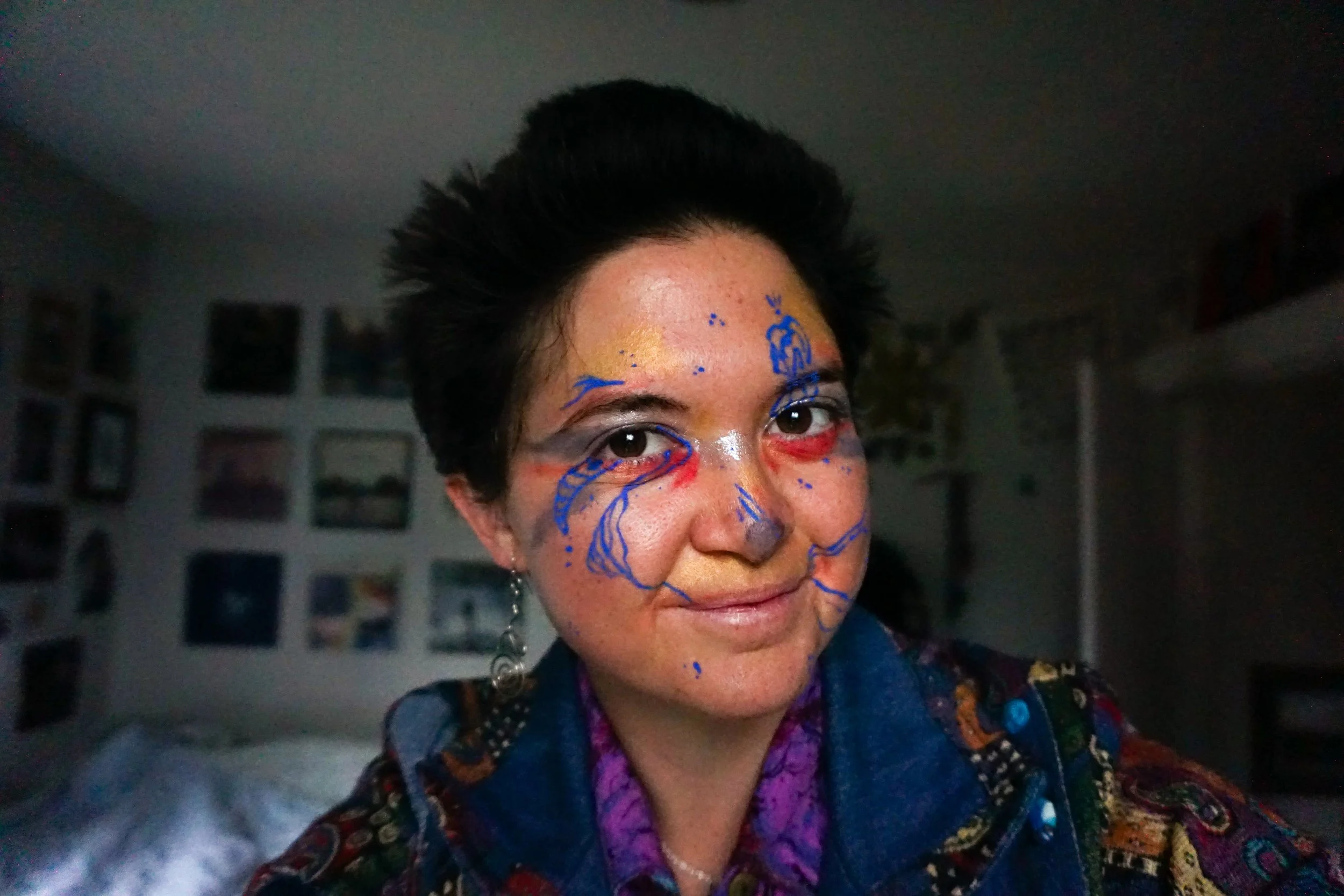 Person with short dark hair, wearing colorful clothing, and face paint with blue, red, and yellow doodles on their face, smiling slightly.