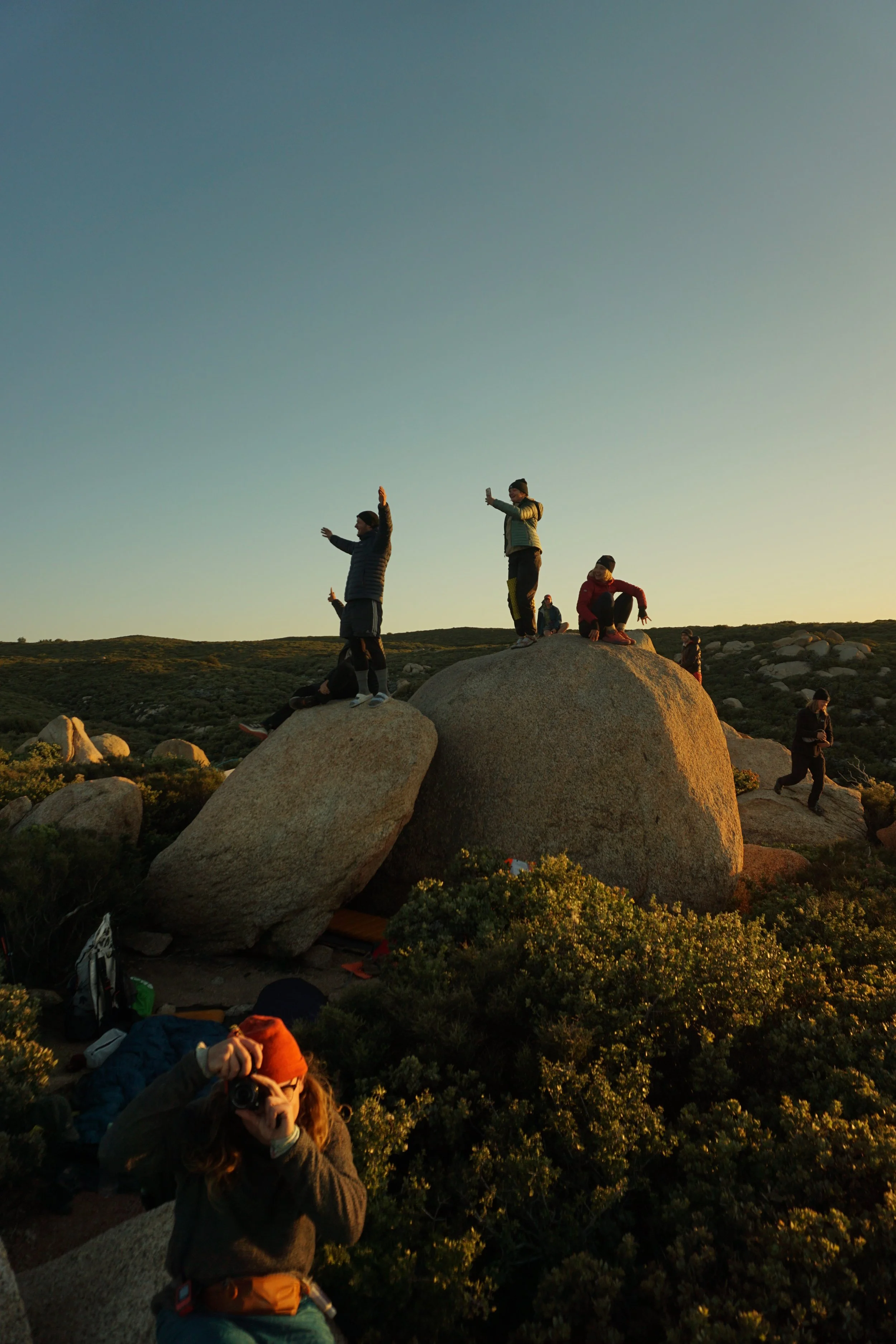 Group of people standing and sitting on large rocks in a landscape at sunset, some taking photos and enjoying the view.