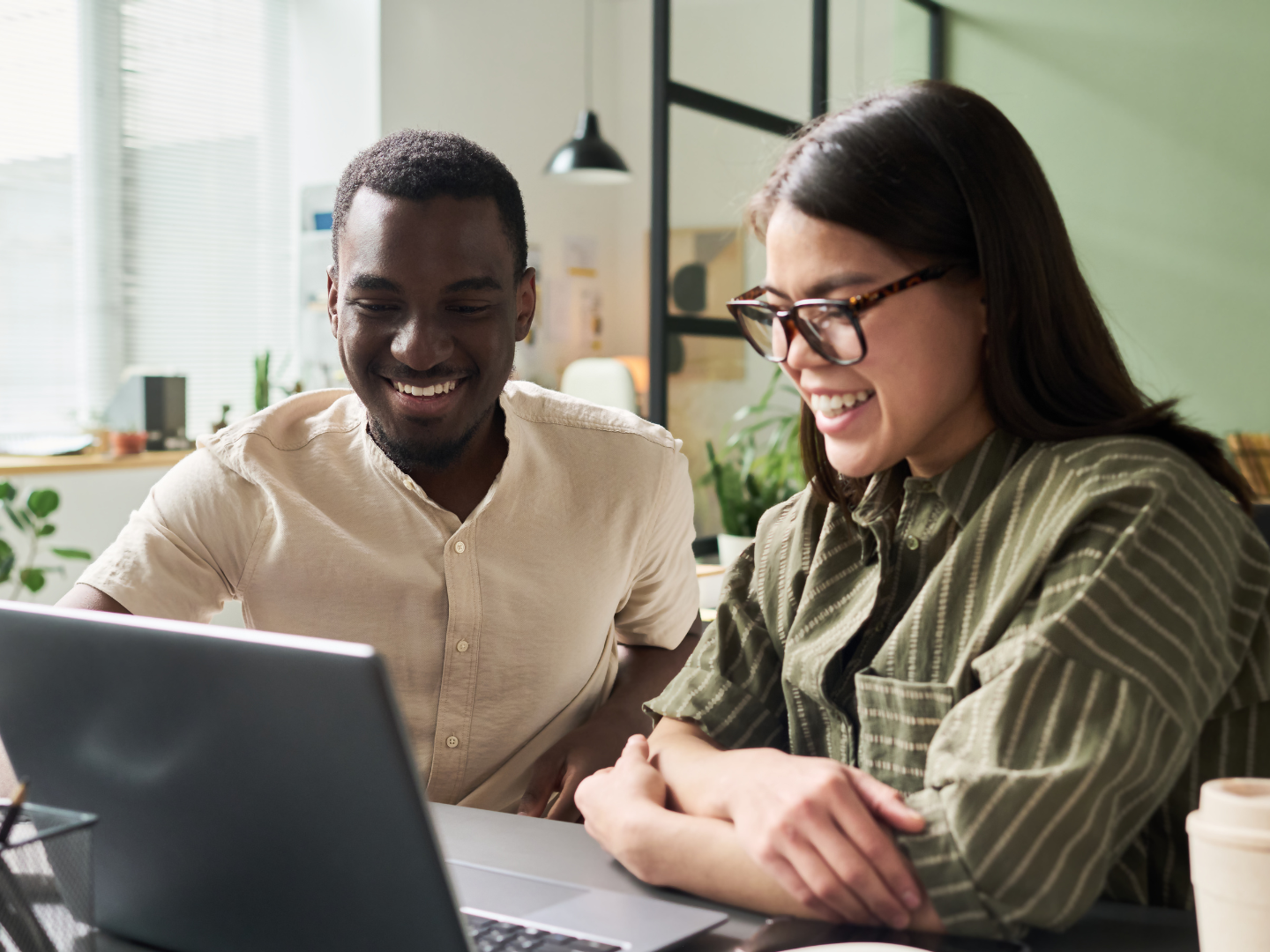 Two people, a man and a woman, smiling while looking at a laptop in a bright office space.