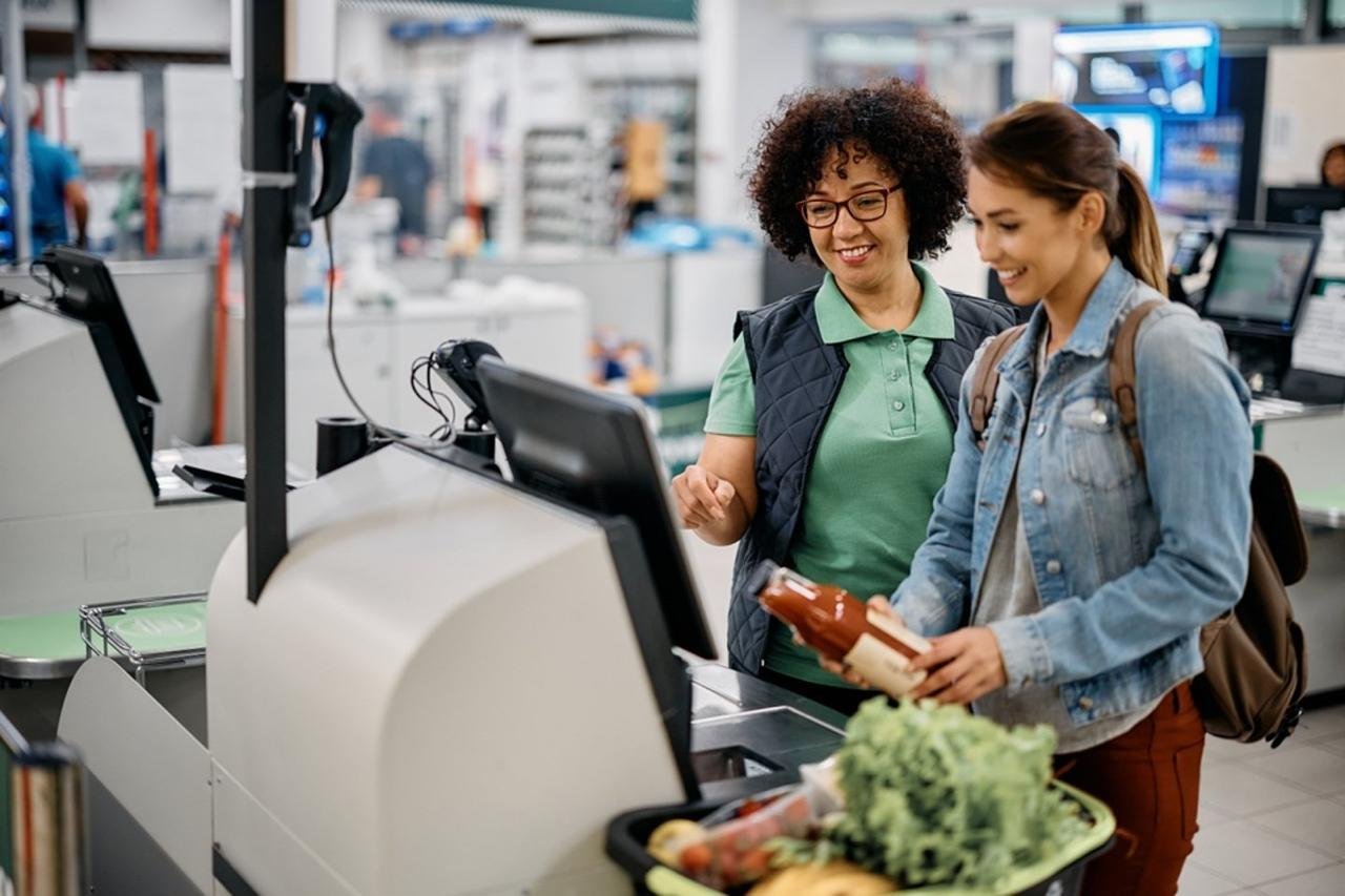Two women shopping at a grocery store checkout counter, with one holding a bottle of sauce and a shopping basket of vegetables in front of them.