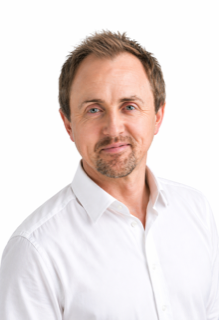 Portrait of a man with light brown hair, a beard, wearing a white shirt, against a white background.
