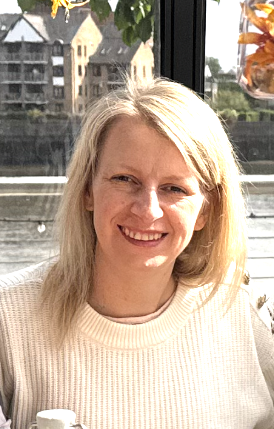 A smiling woman with blonde hair sitting near a window with a river and residential buildings outside.