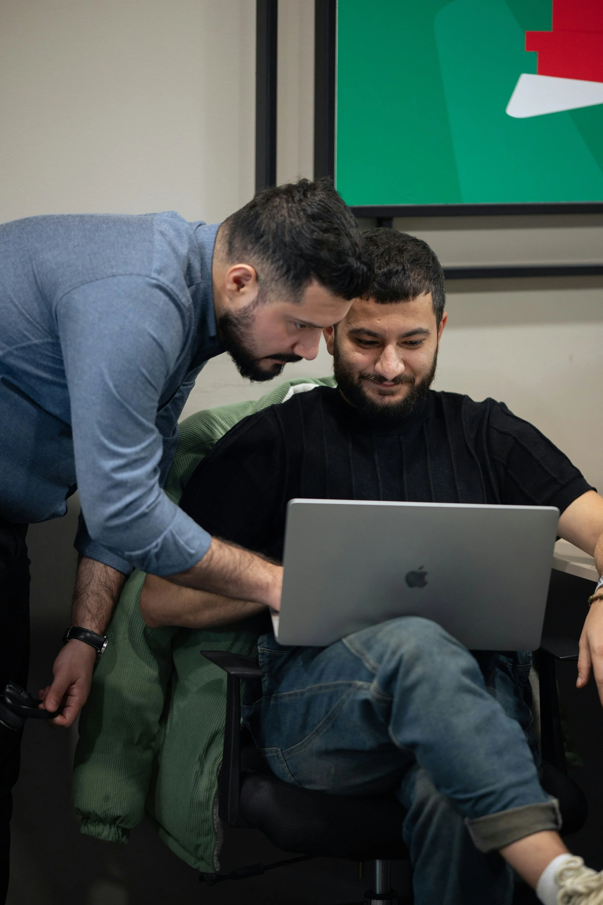 Two men are looking at a laptop, with one leaning over the other who is sitting with legs crossed and wearing a green jacket and jeans. A large screen with a colorful graphic is mounted on the wall behind them.