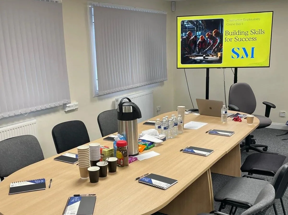 Meeting room with a large table, chairs, a laptop, bottled water, snacks, notebooks, and a large screen displaying a presentation titled 'Building Skills for Success' for a construction employability course.