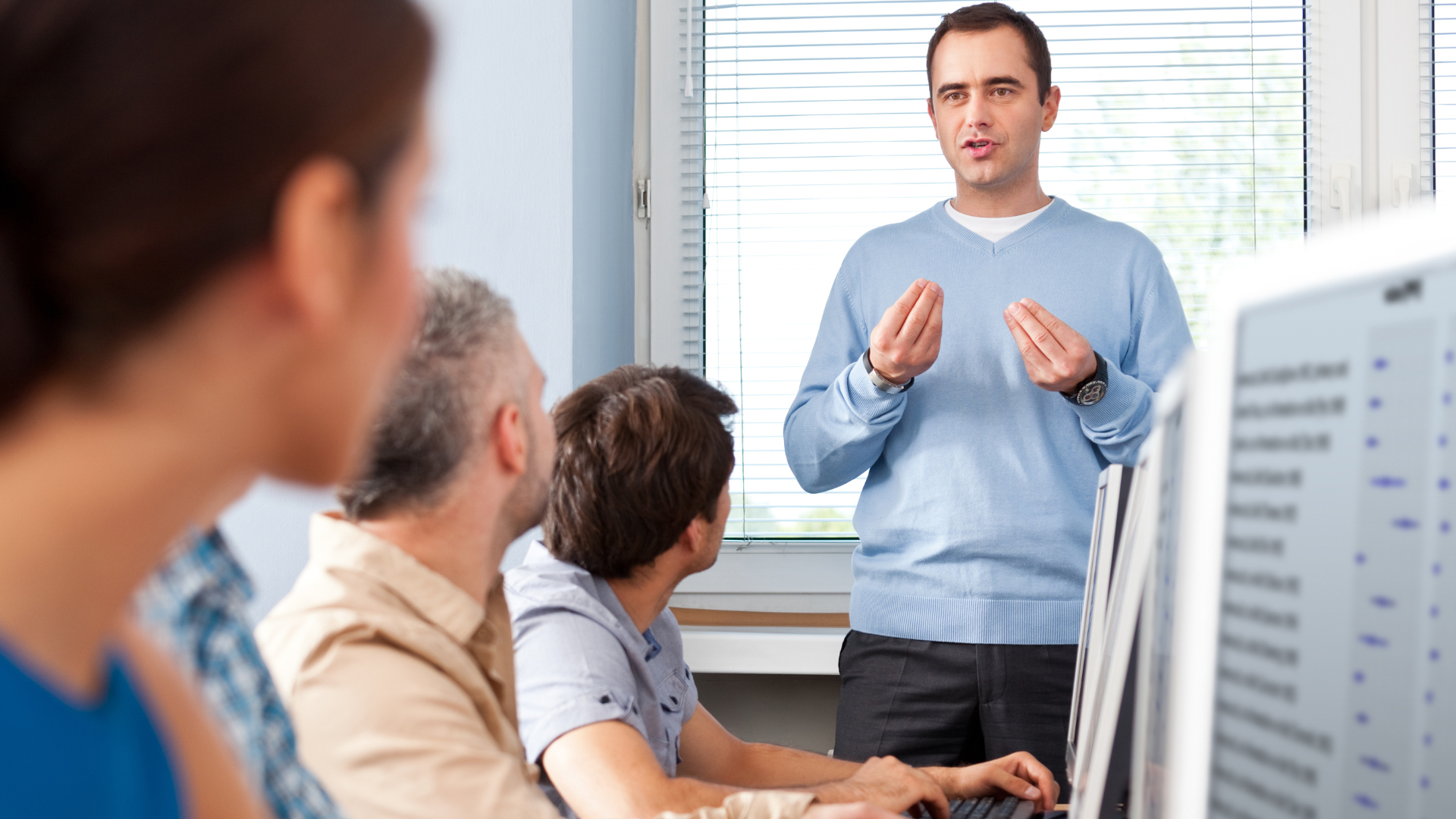 A man in a blue sweater giving a presentation to a group of four seated people in an office with large windows and blinds.