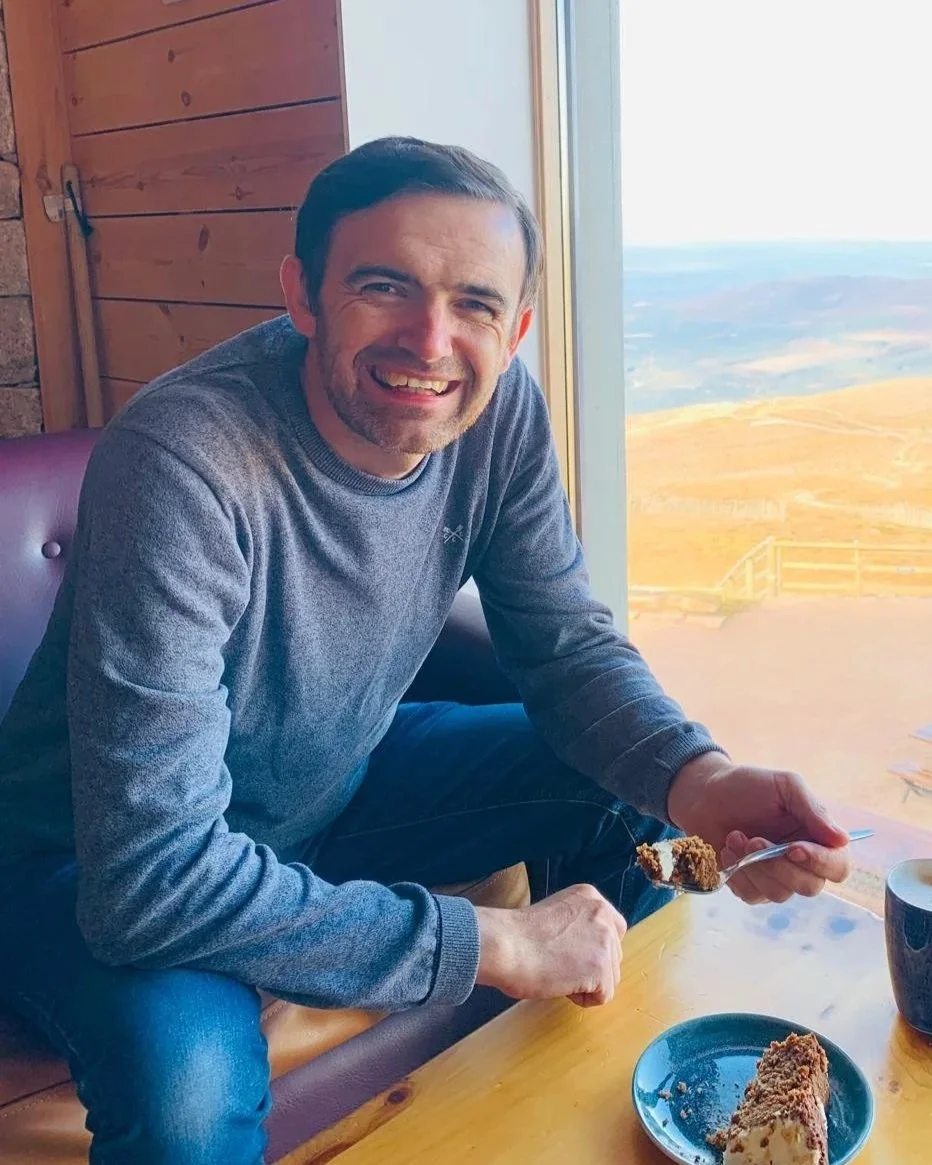 A man smiling while sitting at a wooden table near a window.