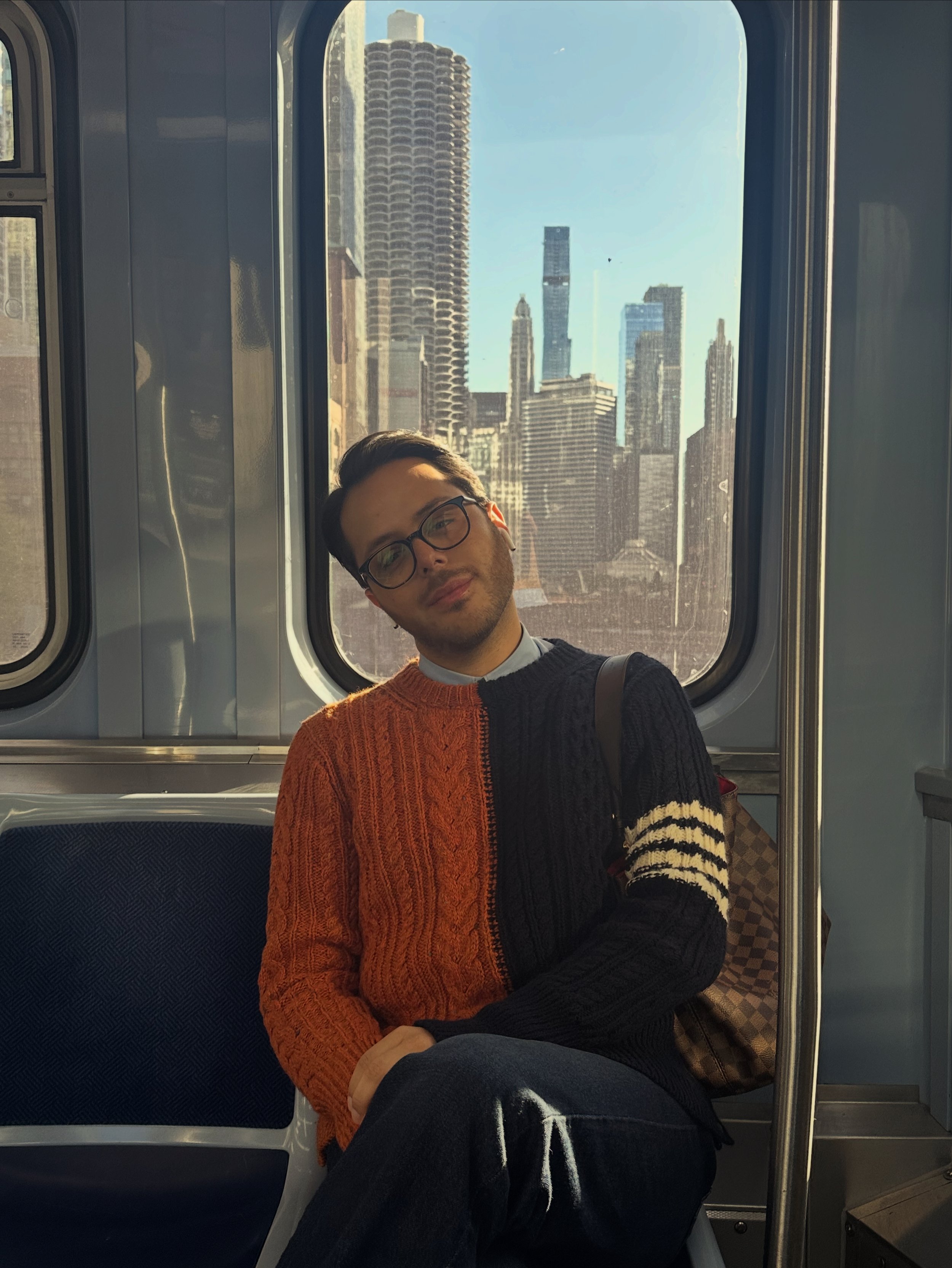 A young man with glasses sitting on a subway train, wearing a multicolored sweater with orange, black, and white stripes, with a cityscape visible through the window behind him.