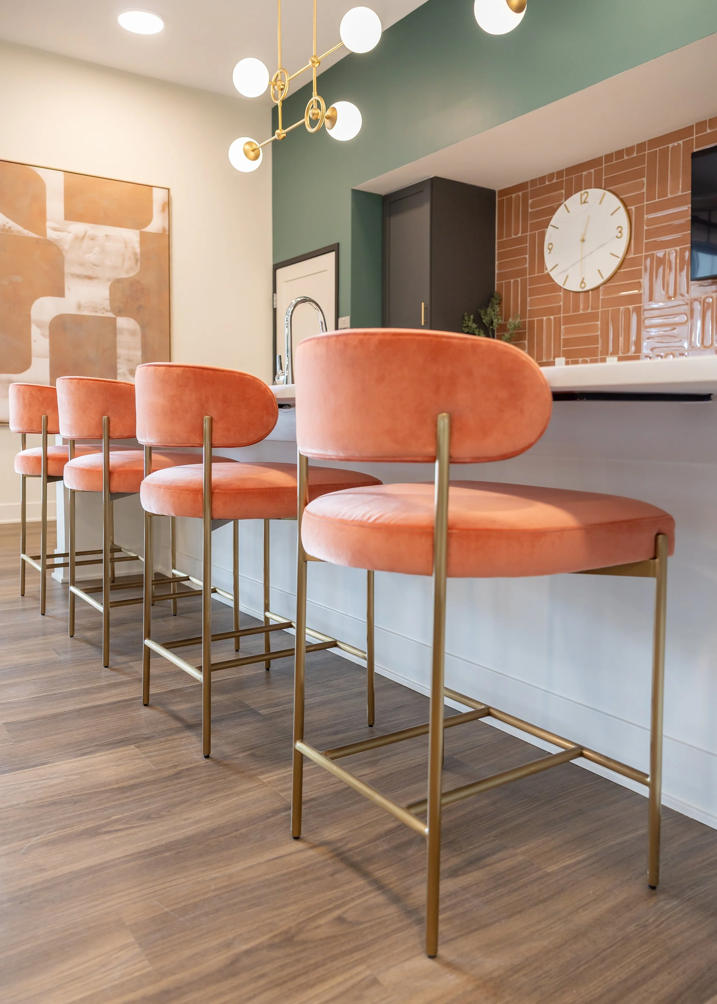 Modern kitchen interior with a row of coral velvet barstools with gold legs lined up at a white kitchen island. A hanging light fixture with round bulbs and gold accents is visible, along with a wall clock, a mounted TV, a plant, and abstract artwork on the wall.