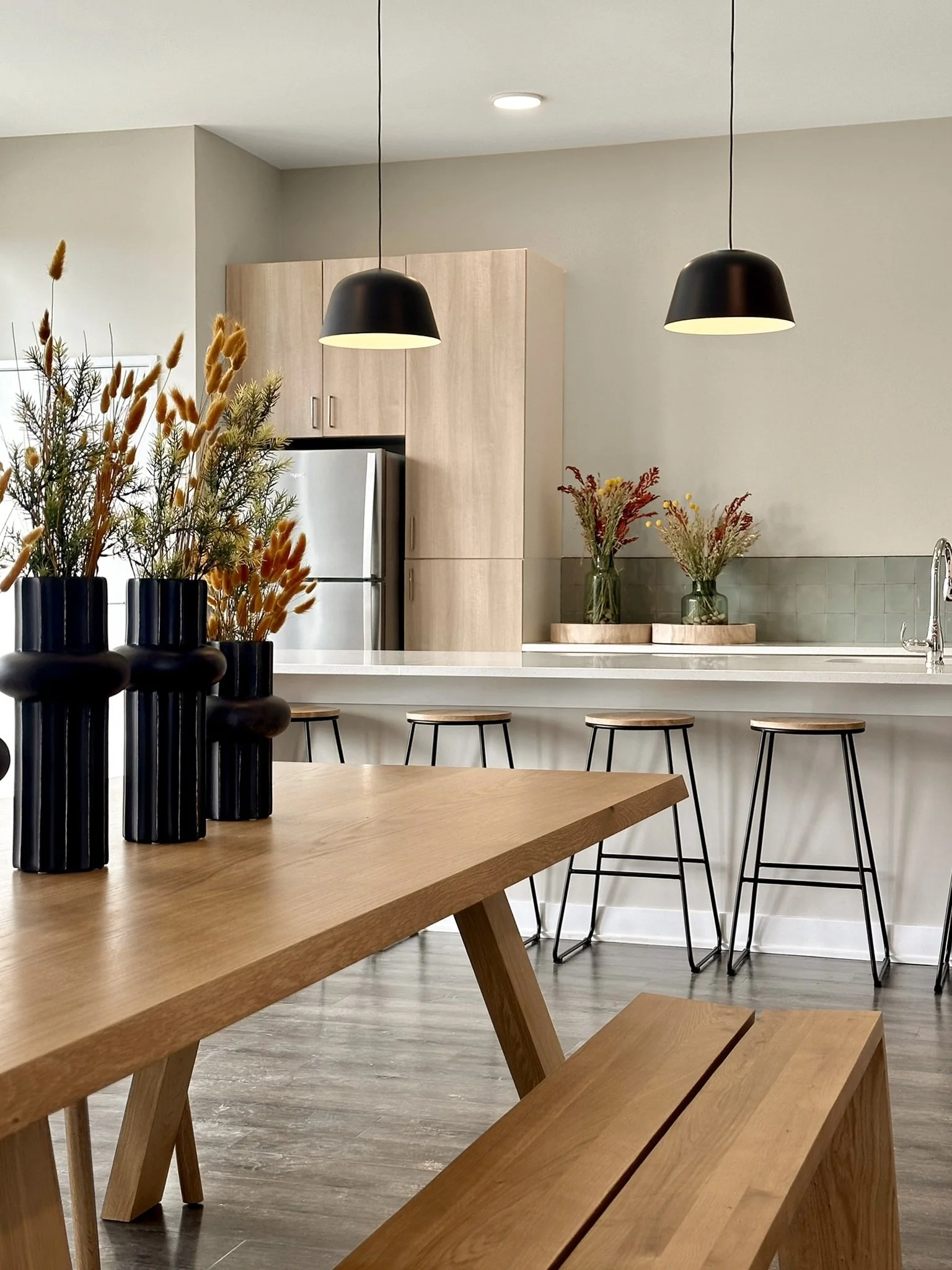 A modern kitchen with a wooden dining table and matching benches, three black vases with dried flowers, a kitchen counter with three wooden barstools, a kitchen cabinet, and two black pendant lights hanging from the ceiling.