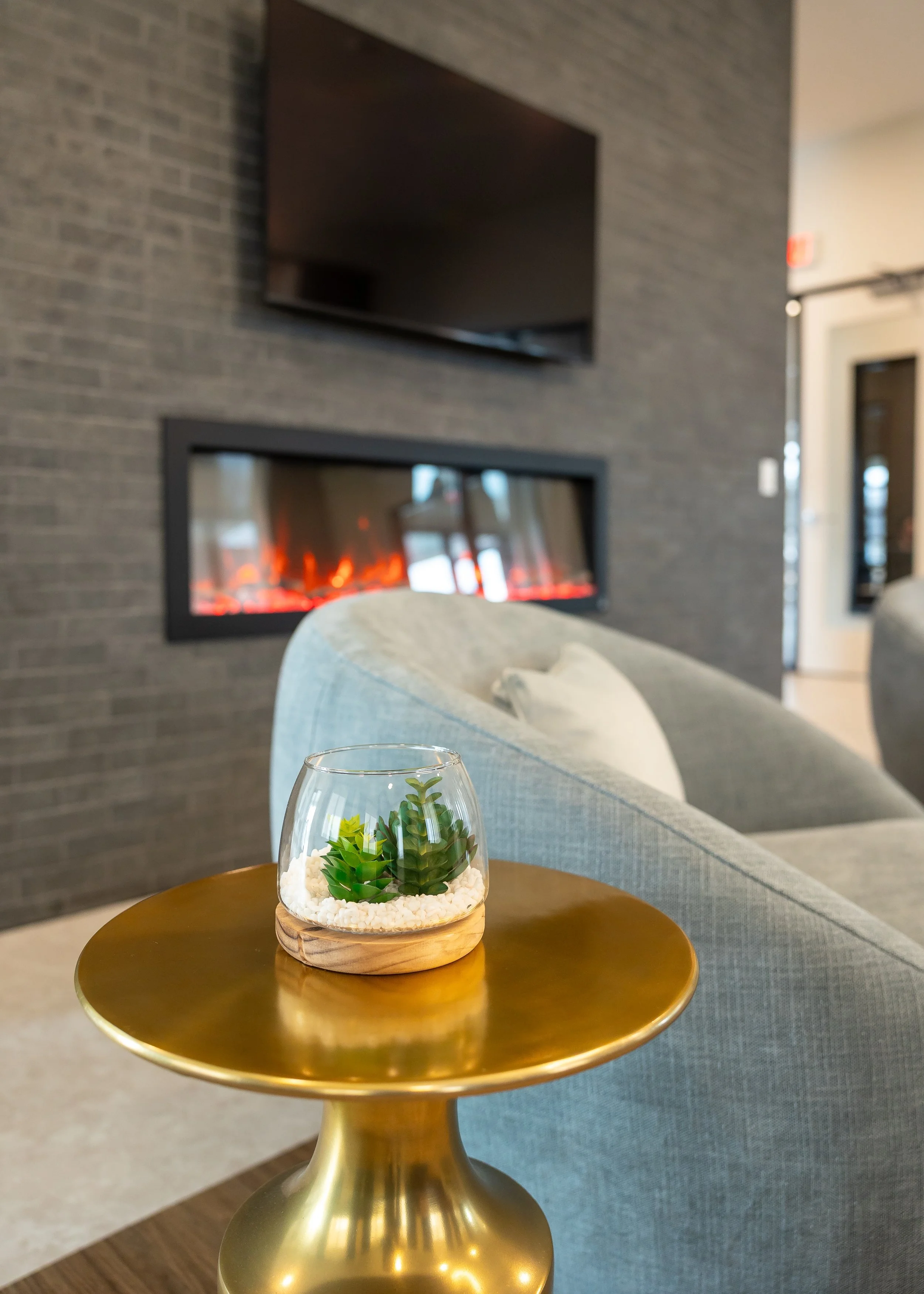 A small glass terrarium with succulents and white stones on a gold-colored side table in a modern living room with a brick wall, a mounted TV, and a fireplace.