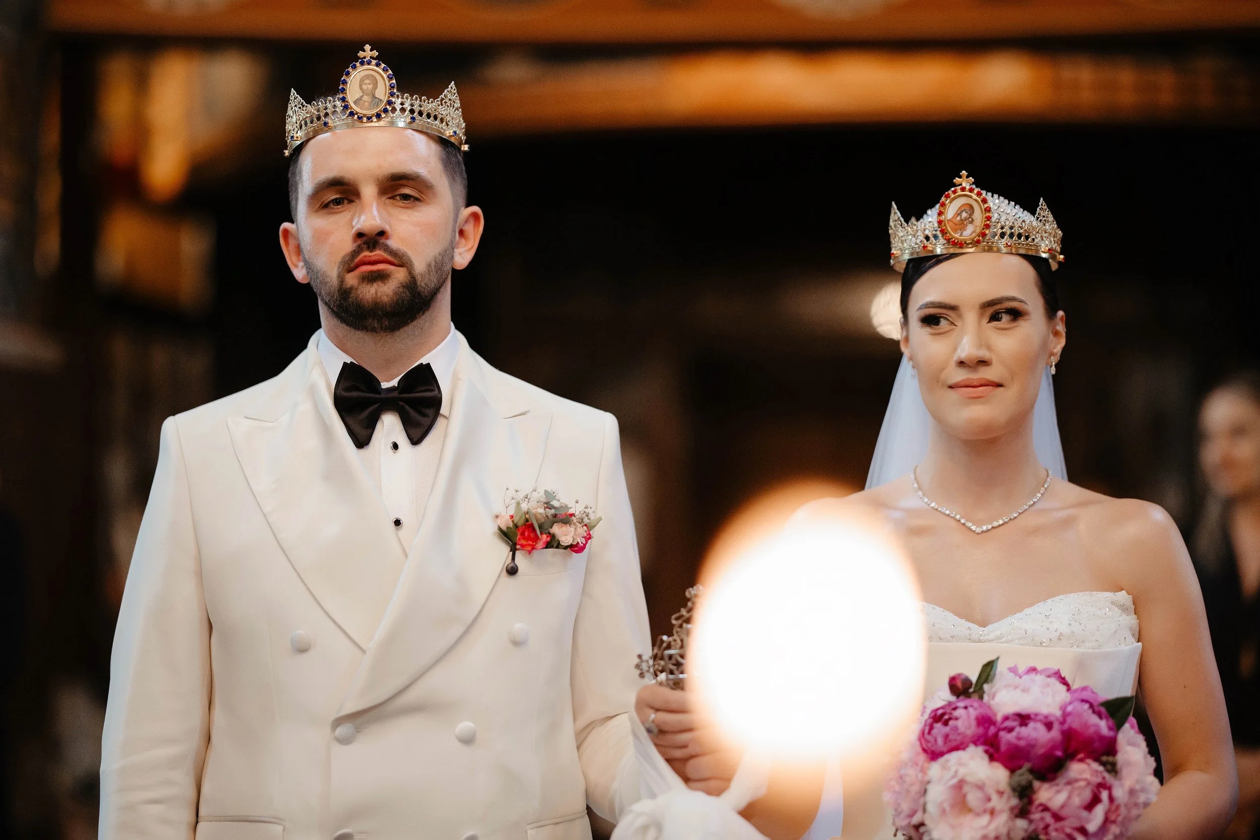 A bride and groom wearing crowns during their wedding ceremony. The groom is in a white tuxedo with a black bow tie, holding a bouquet of pink flowers. The bride is in a strapless white wedding dress with a pearl necklace, holding a bouquet of pink a
