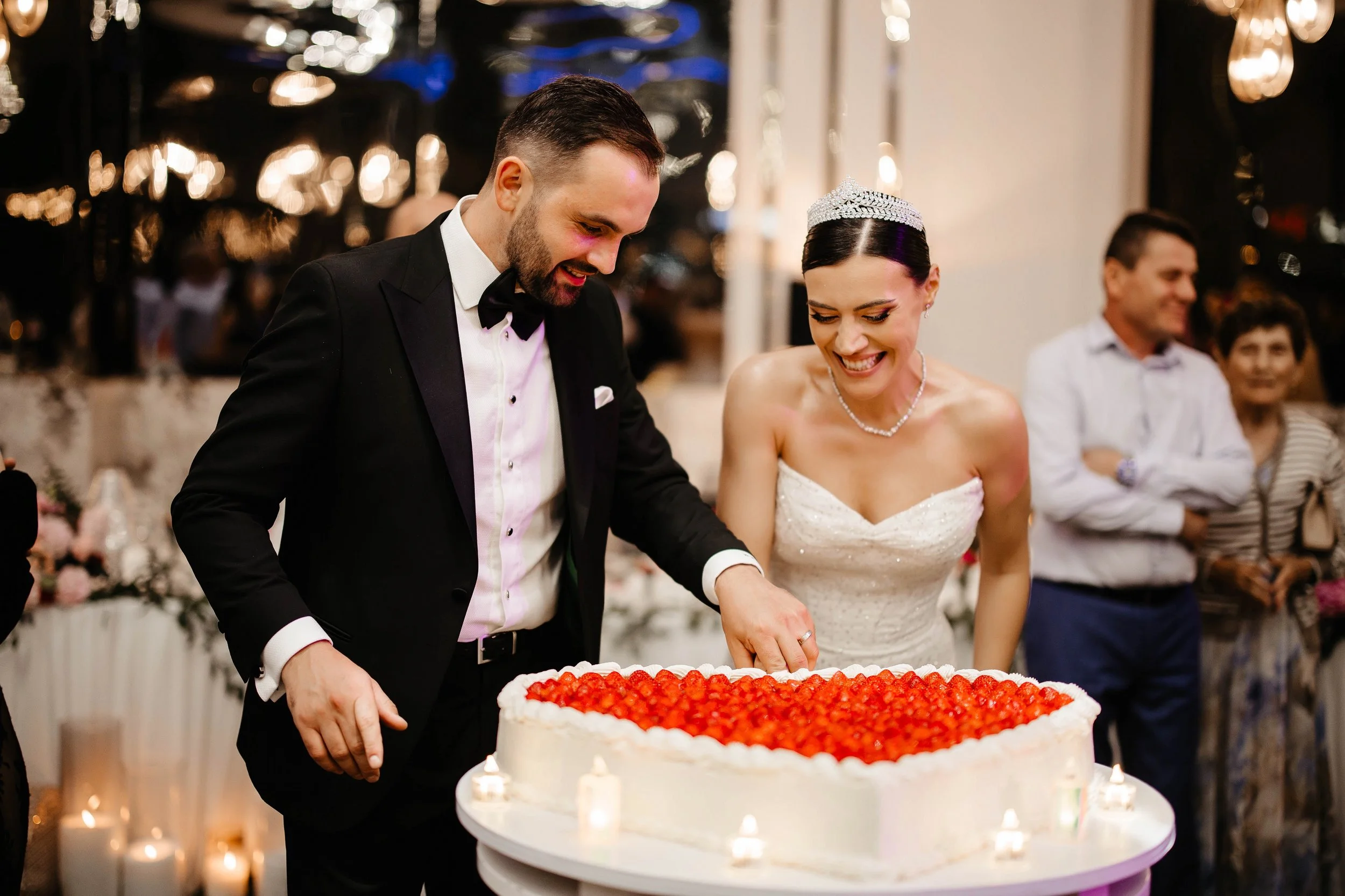 A newlywed couple and guests cutting a wedding cake during a celebration, with the bride wearing a tiara and the groom wearing a tuxedo.