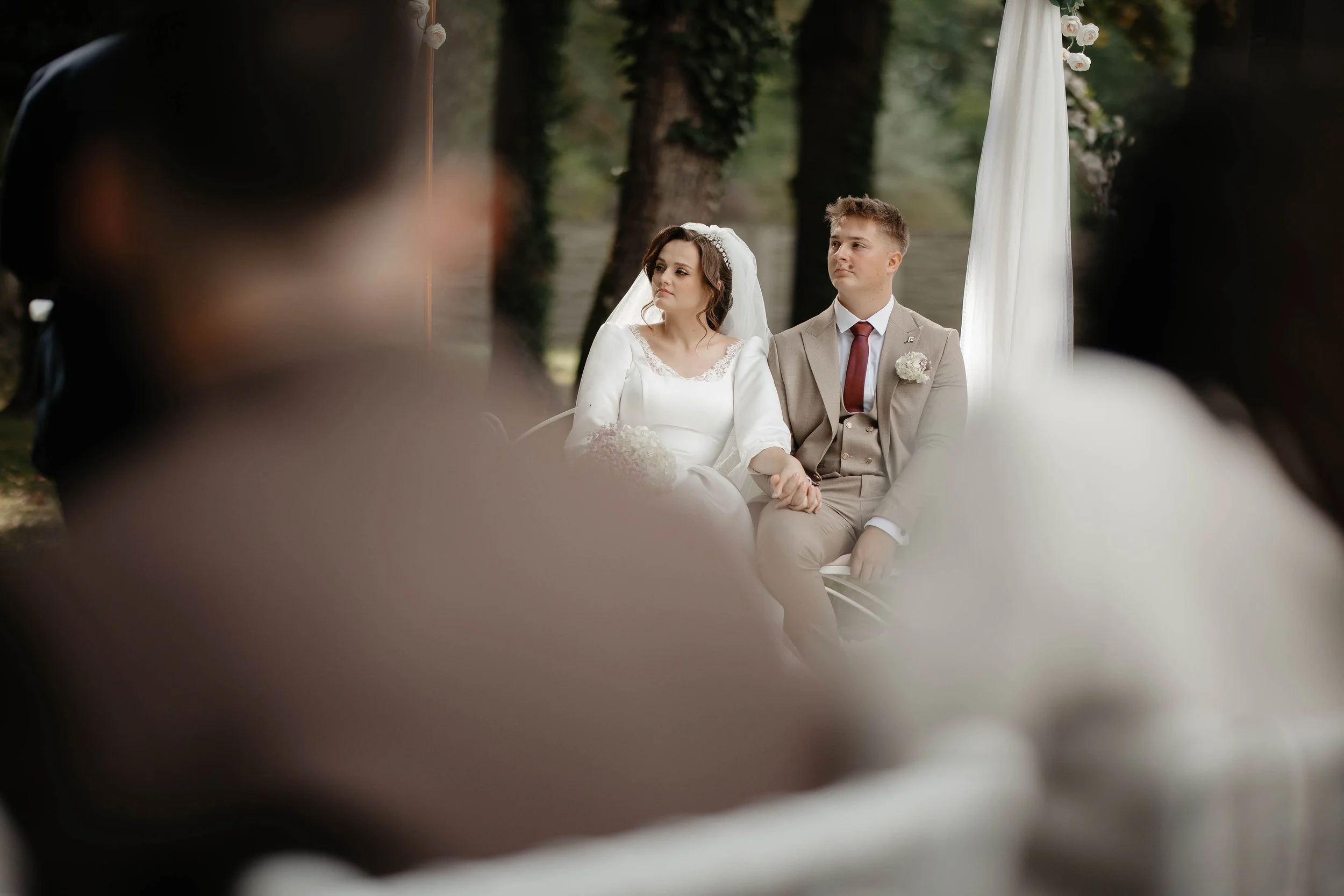 A bride and groom sitting outdoors during their wedding ceremony, looking serious, with trees in the background and a white draped fabric with flowers behind them.
