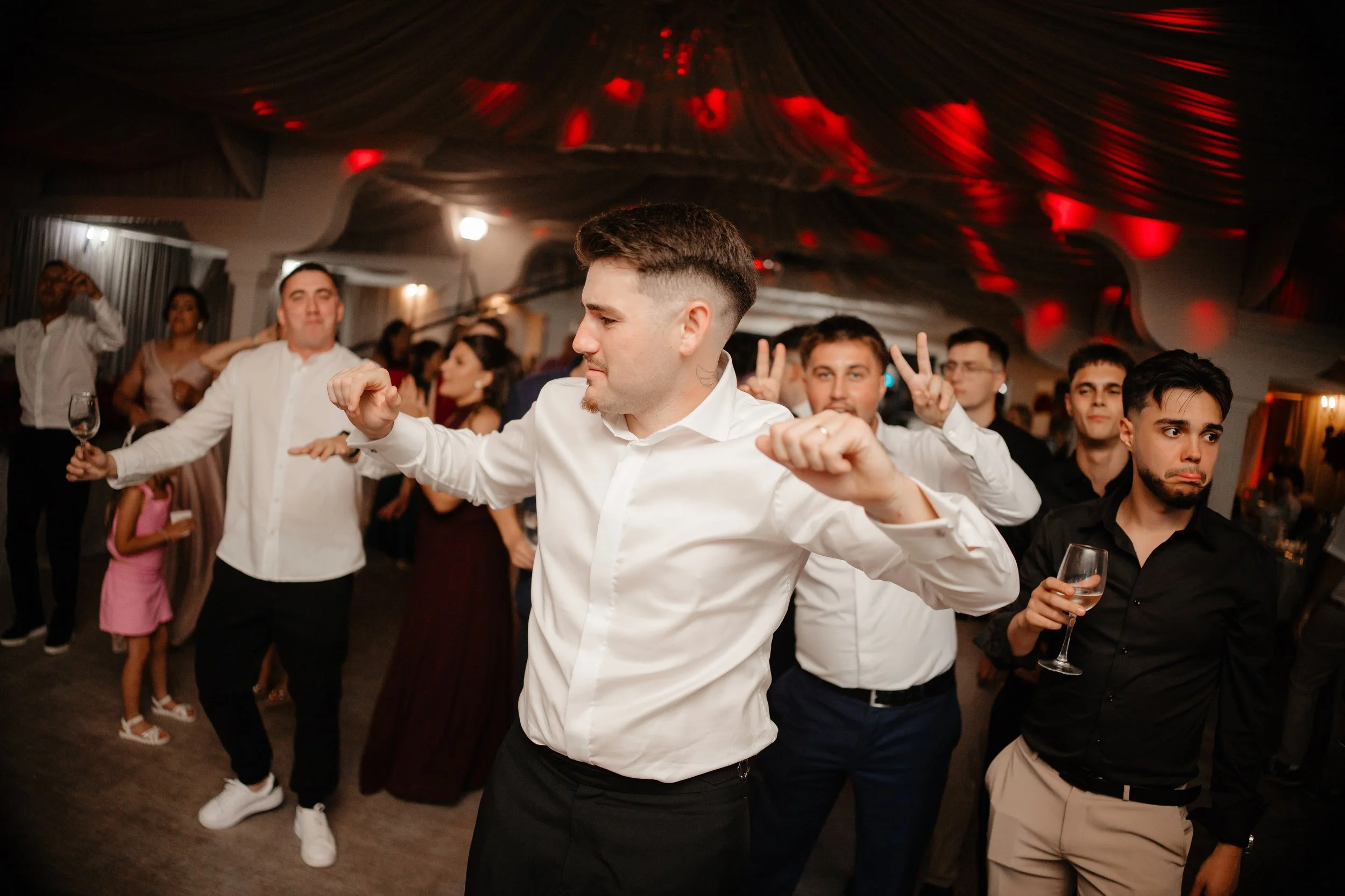 People dancing and socializing at a party or celebration in an indoor venue with dim lighting and red accents on the ceiling.