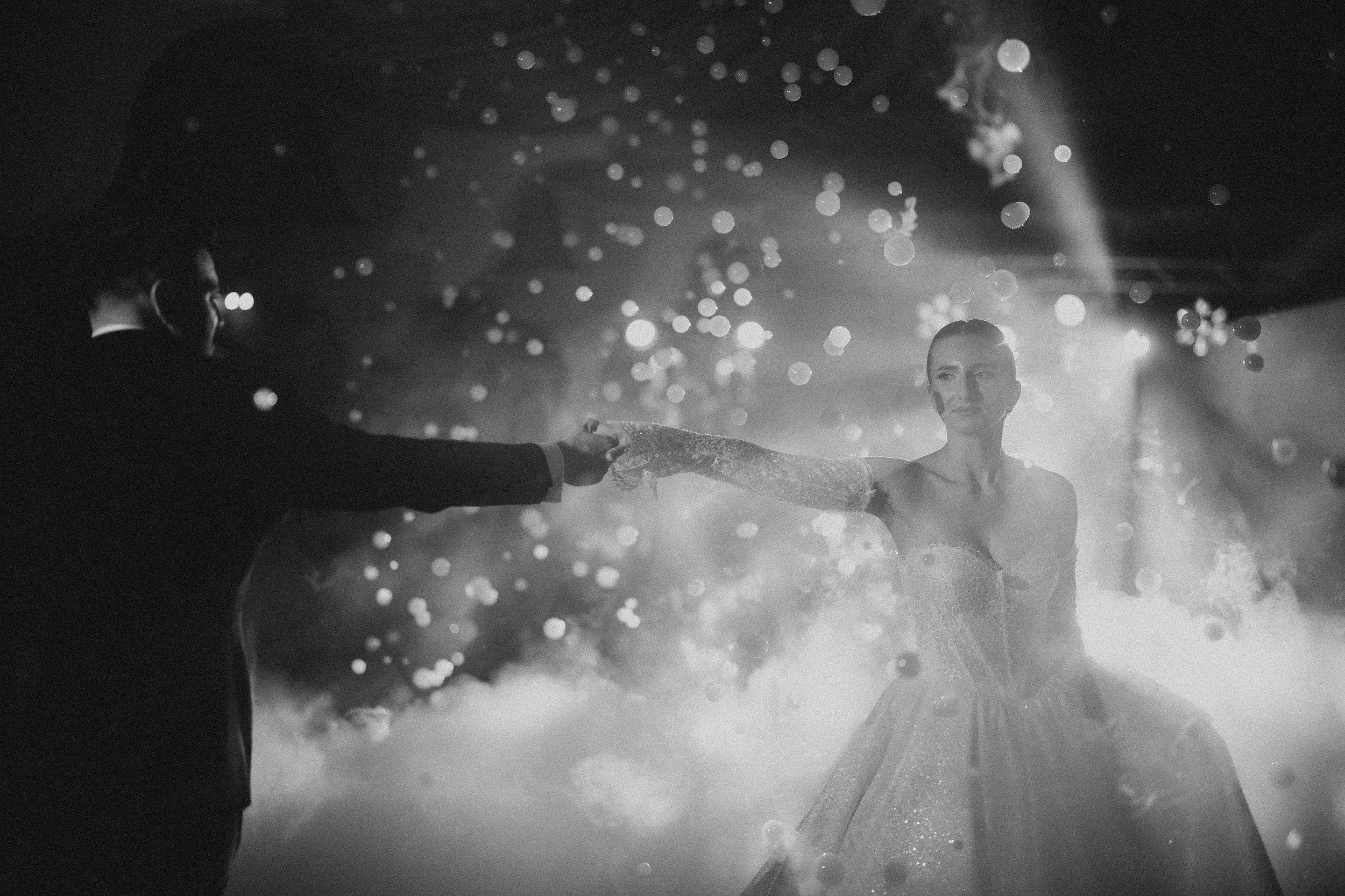 A black and white photo of a couple dancing, with the man wearing a suit and the woman in a sparkling dress, illuminated by stage lights and surrounded by falling confetti.