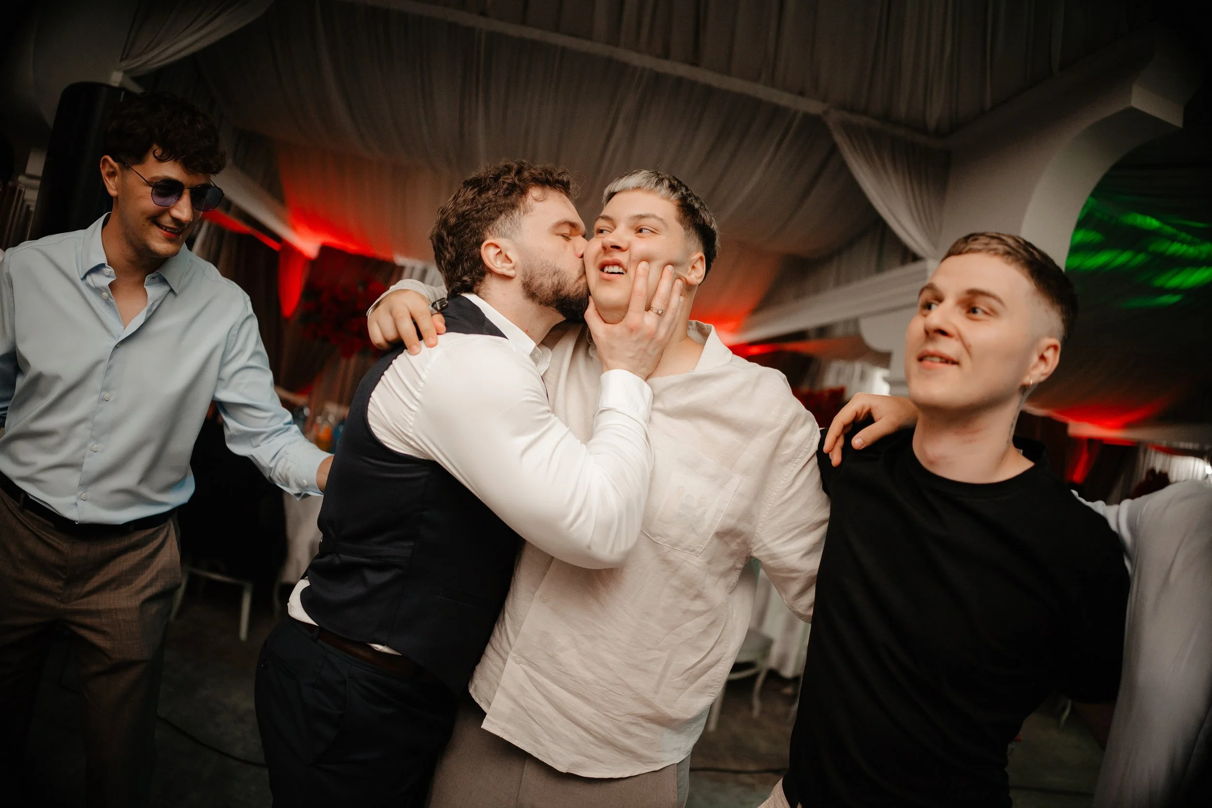 Four young men at a celebration or party, dancing together and enjoying themselves, with one kissing another on the cheek.