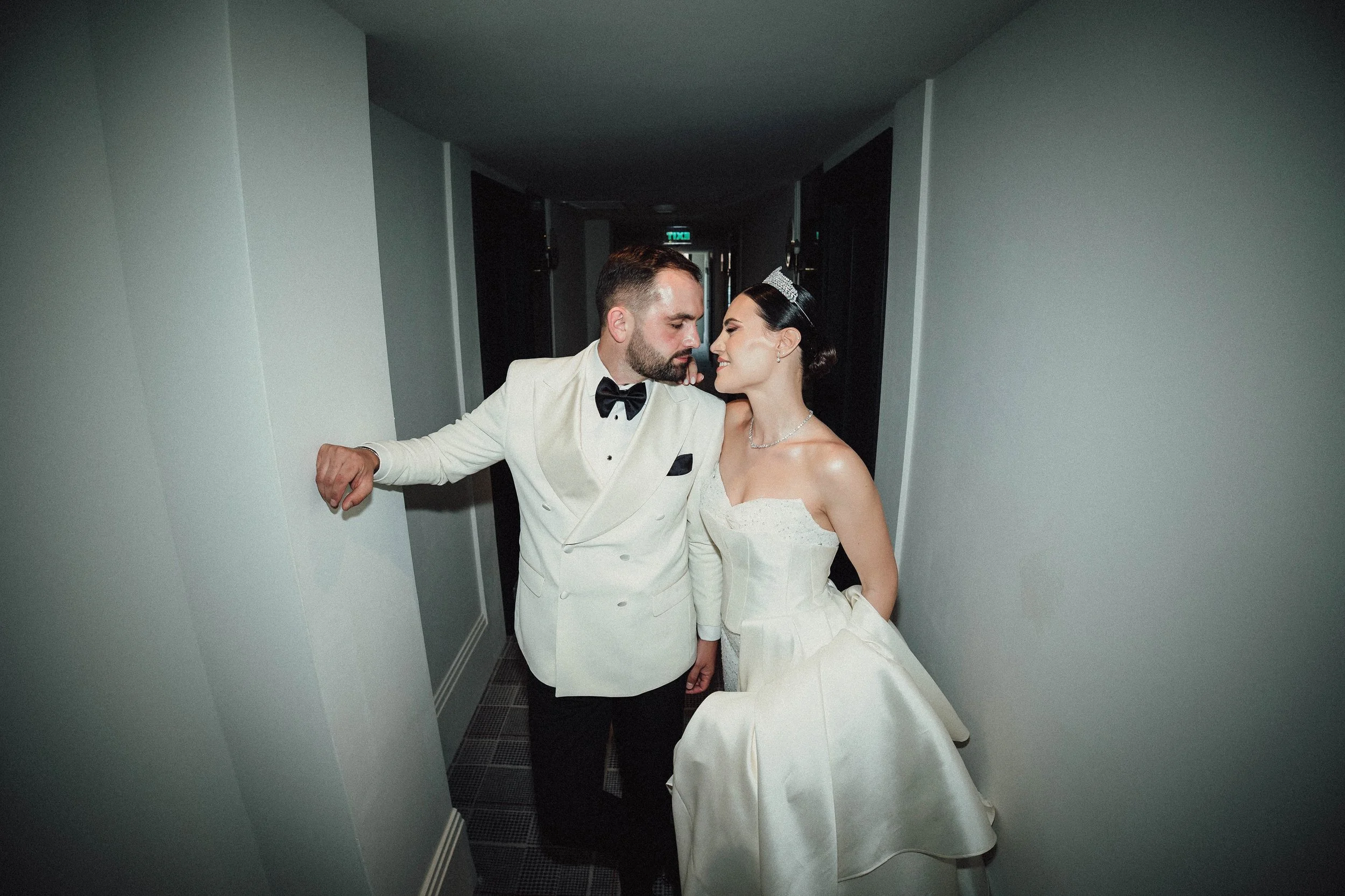 A bride and groom in wedding attire standing very close together in a narrow hallway, with the groom dressed in a white tuxedo and bow tie, and the bride in a strapless wedding dress with a tiara, both leaning towards each other with a romantic expre