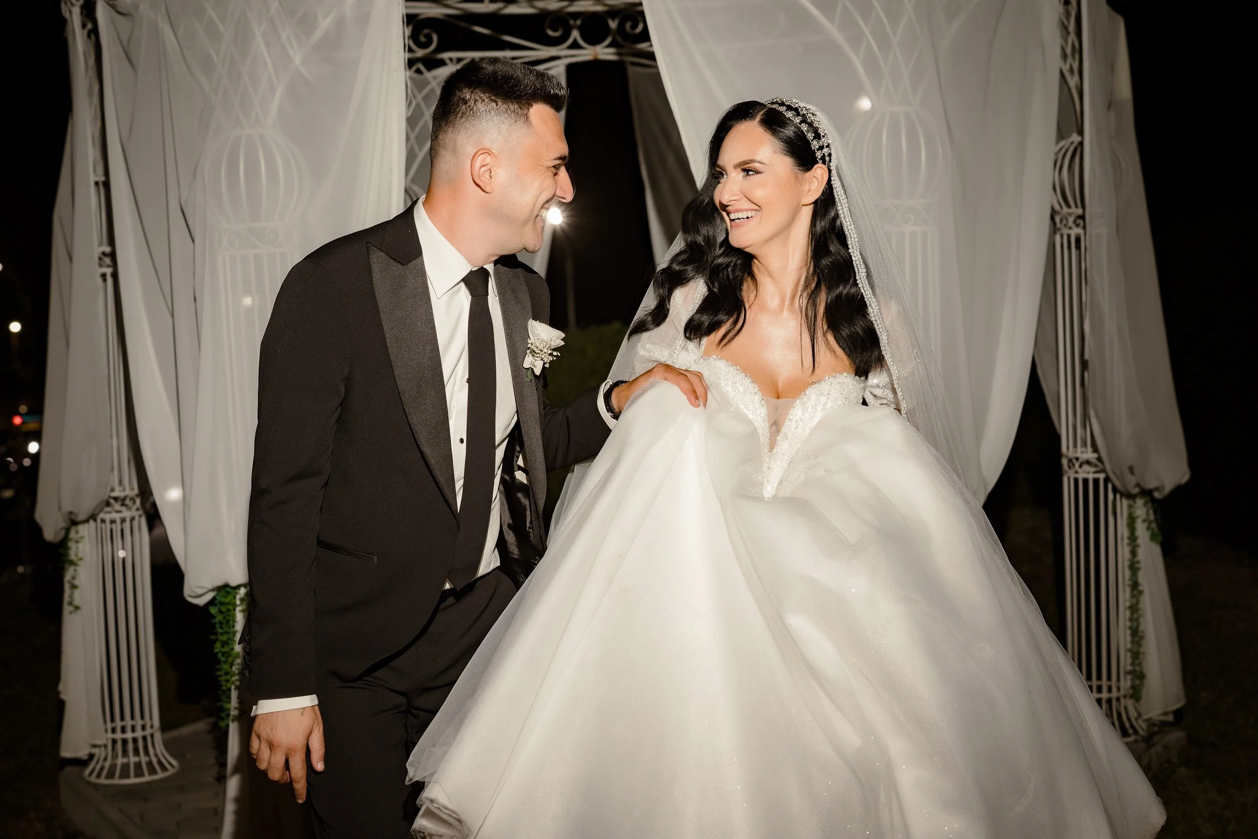 A bride and groom smile at each other during their wedding ceremony at night, standing under a white canopy with decorative fabric and metalwork, dressed in formal wedding attire.