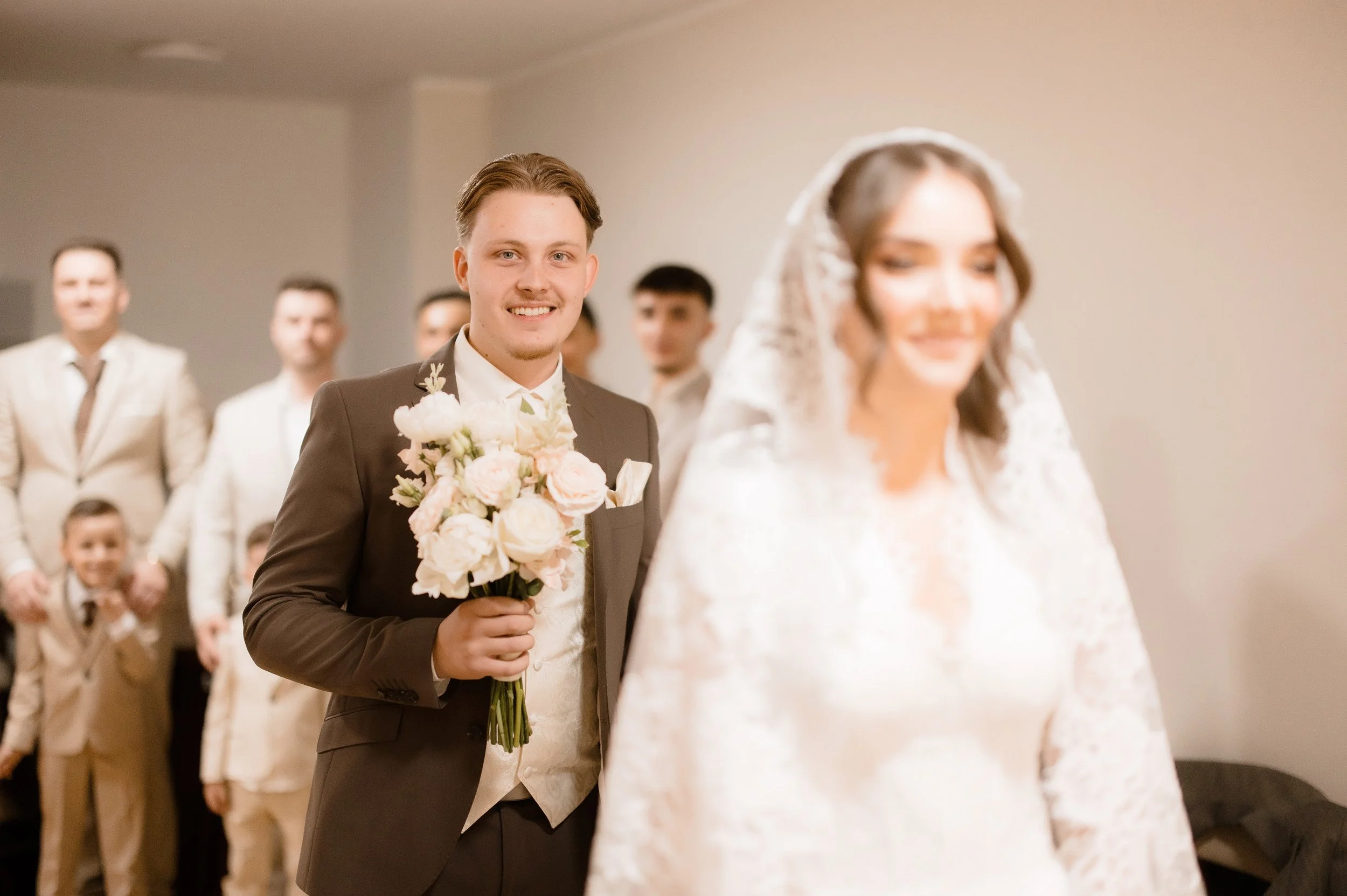 A groom holding a bouquet of pink and white flowers, standing in a wedding ceremony with a smiling bride in the foreground and several groomsmen in beige suits in the background.