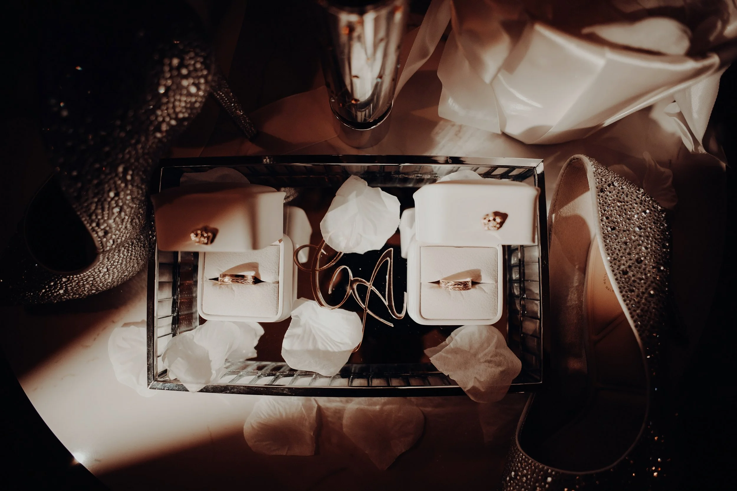 Top view of a jewelry box containing gold rings, with tissue paper and shopping bags around it, accompanied by a glittery purse and high-heeled shoes.