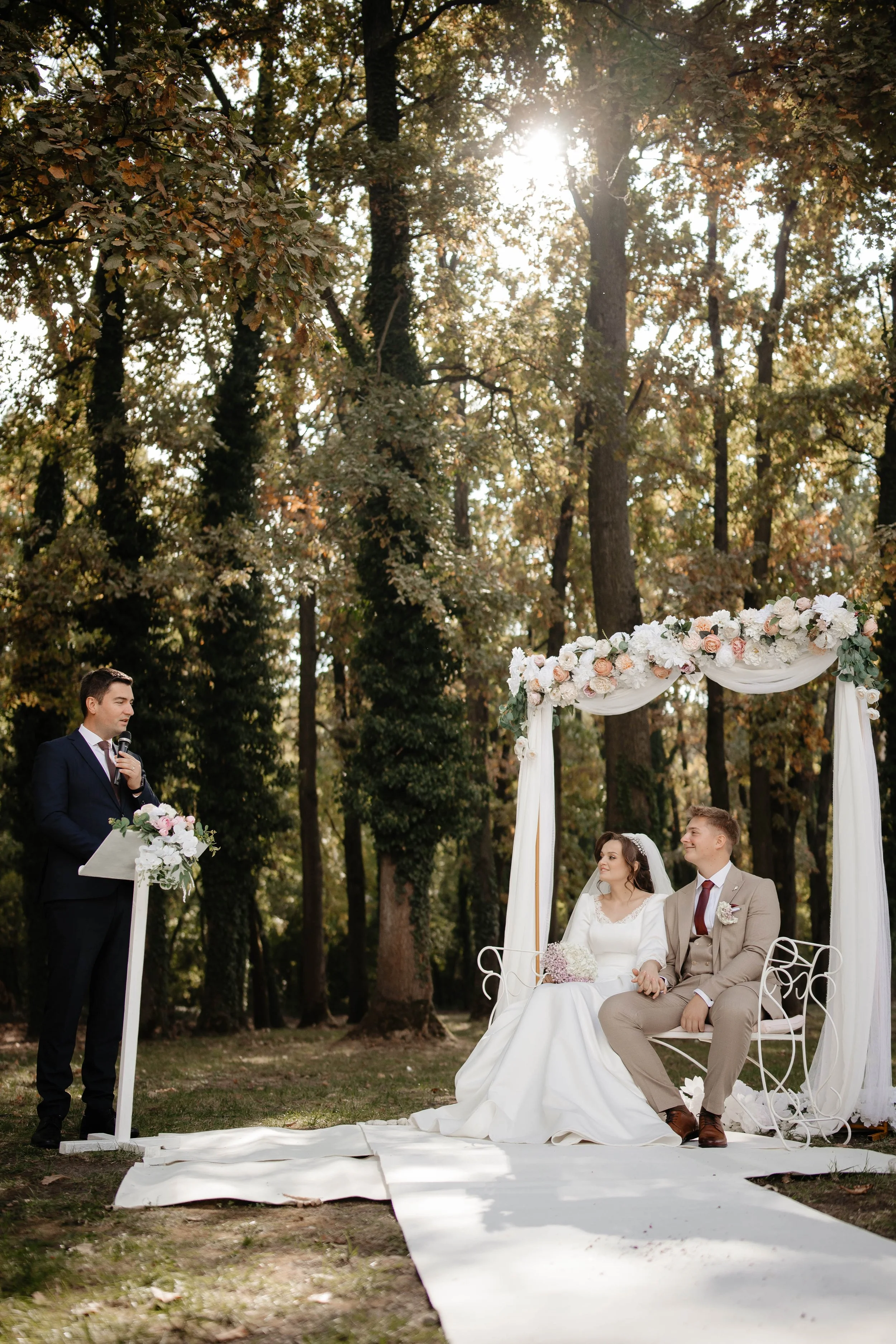 A wedding ceremony in a forest, with a bride and groom sitting under an arch decorated with white and blush flowers, a woman officiant reading from a paper, and a man in a suit standing nearby. Sunlight filters through the trees.