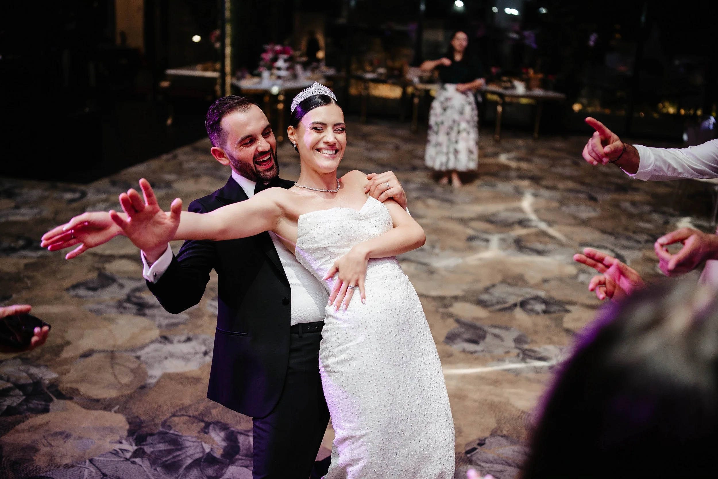 A bride and groom dancing at their wedding reception, with the bride wearing a white wedding dress and tiara and the groom dressed in a black tuxedo. They are smiling and surrounded by people celebrating.