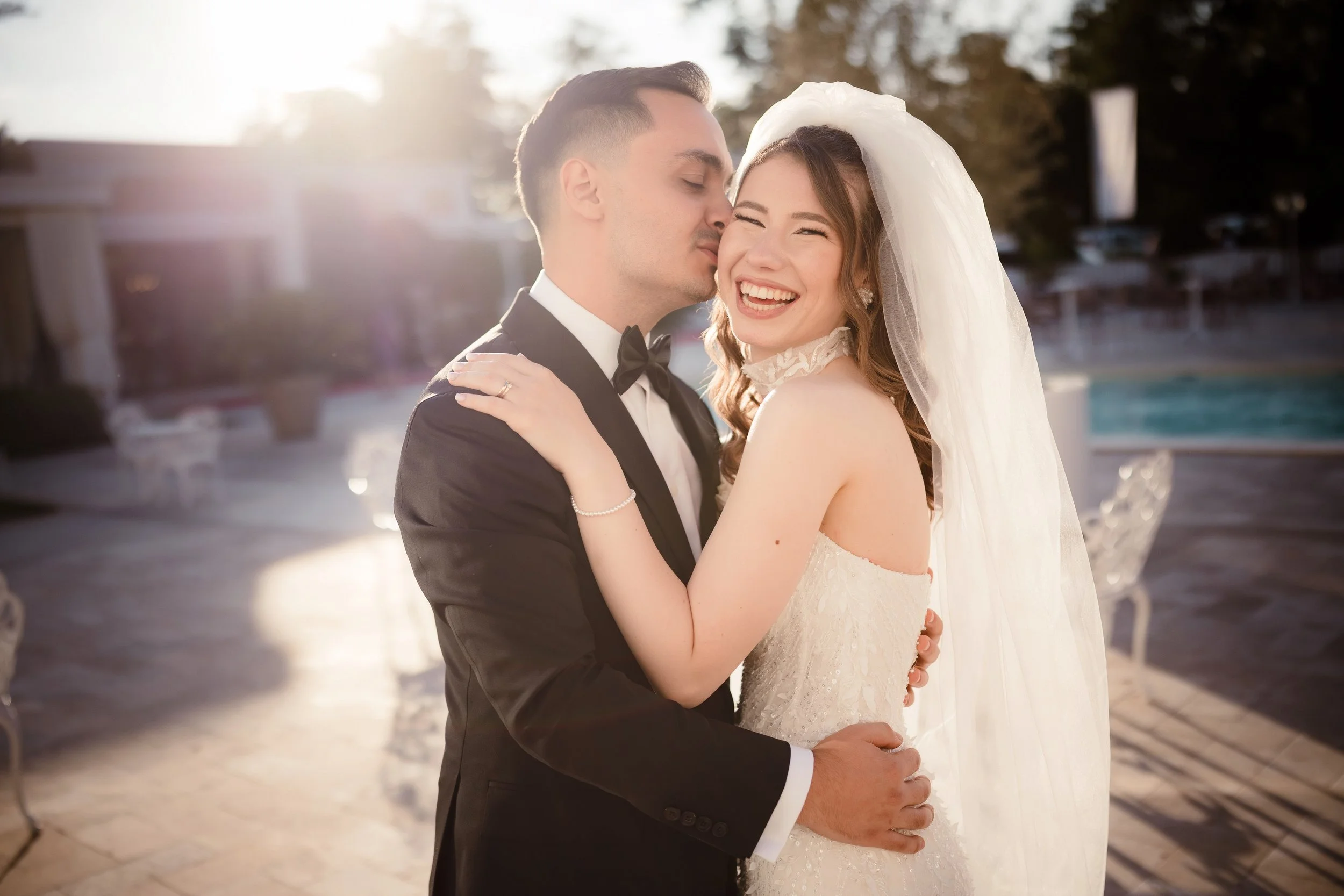 A bride and groom sharing a kiss outdoors, with the bride smiling and wearing a wedding dress and veil, and the groom in a tuxedo, during sunset.