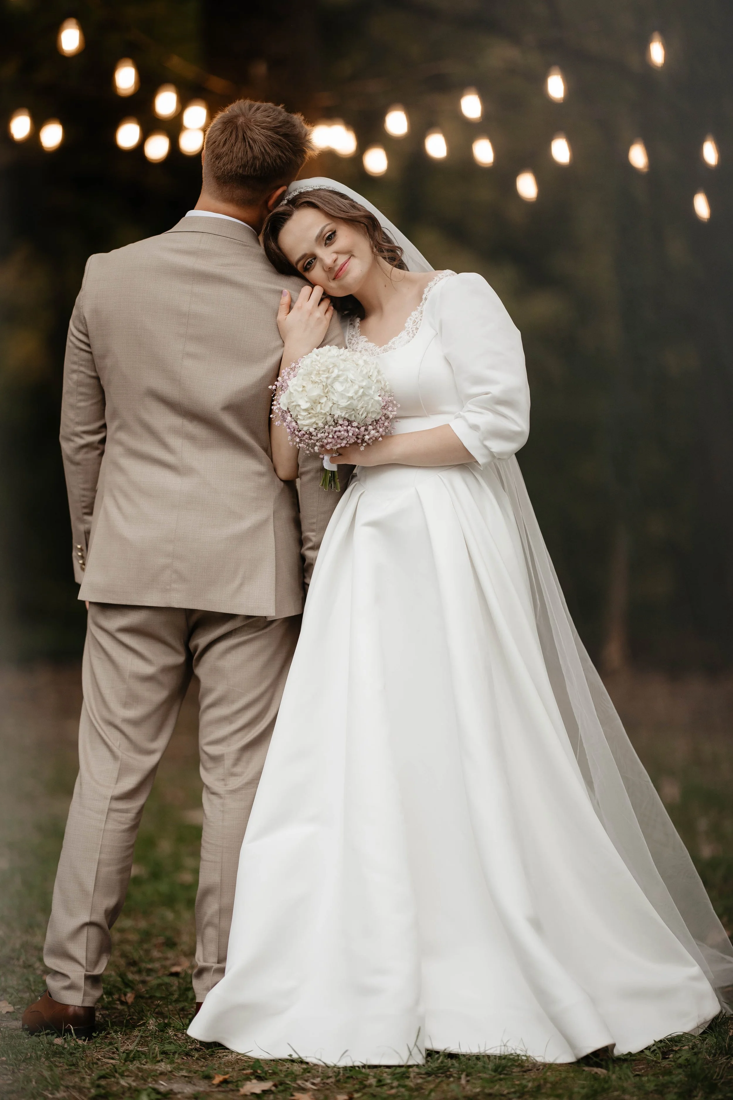 A bride and groom standing outdoors at night, with string lights hanging above, embracing each other. The bride is holding a bouquet and wearing a white wedding dress, leaning her head on the groom's shoulder.