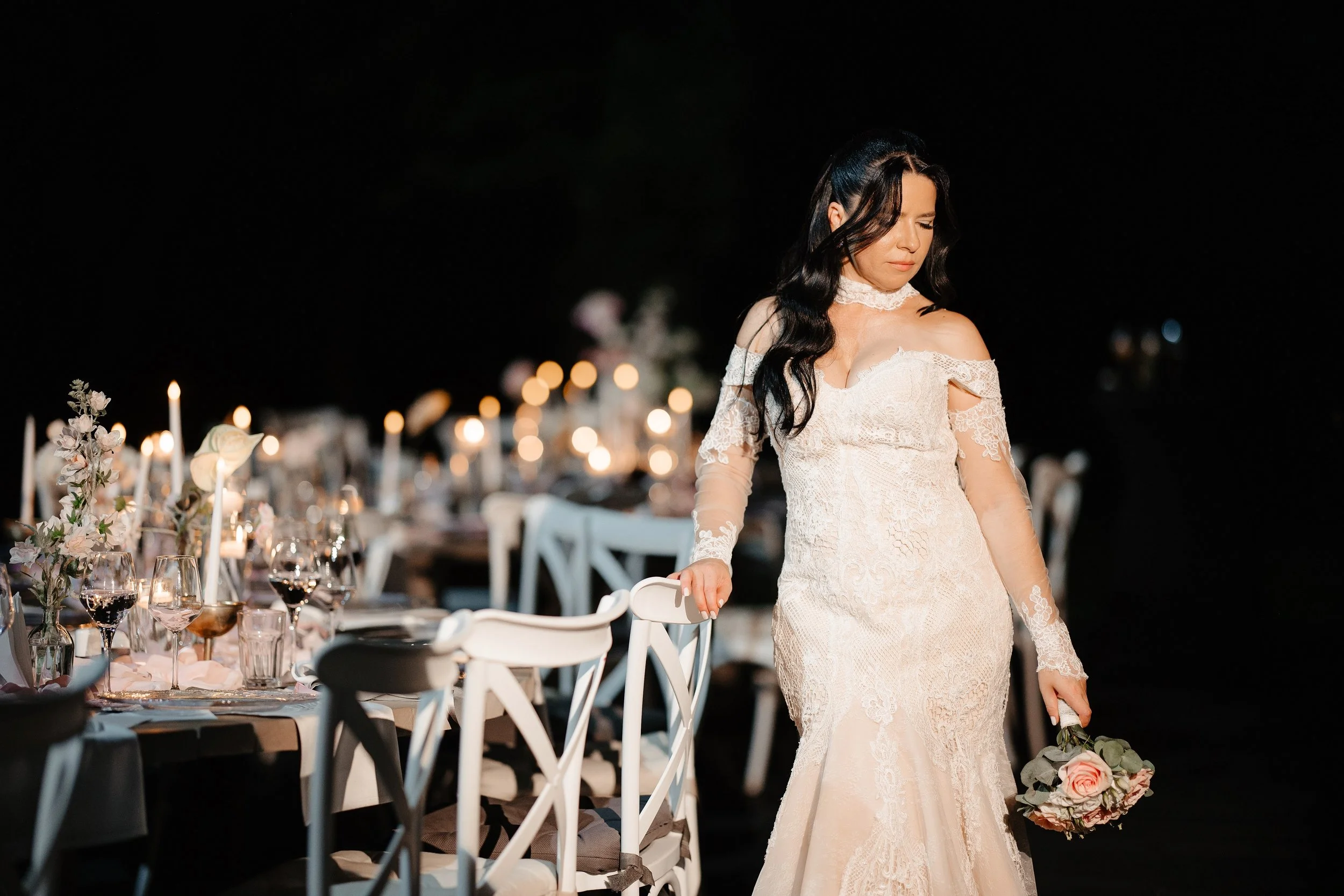 A woman in an off-shoulder lace wedding dress holding a bouquet of pink roses and gray eucalyptus, standing next to a white chair at a nighttime event with decorated tables and candles.