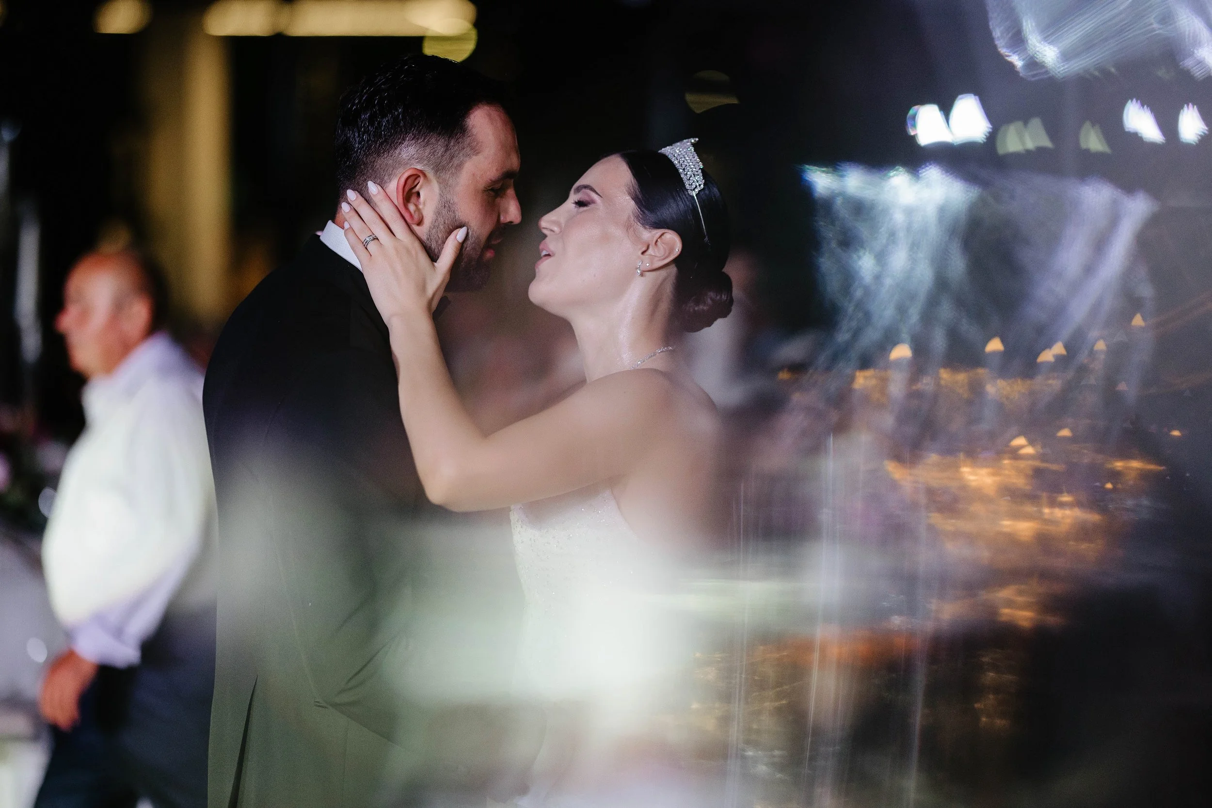 A bride and groom are dancing closely during their wedding reception, with the bride holding the groom's face and smiling while he gazes at her