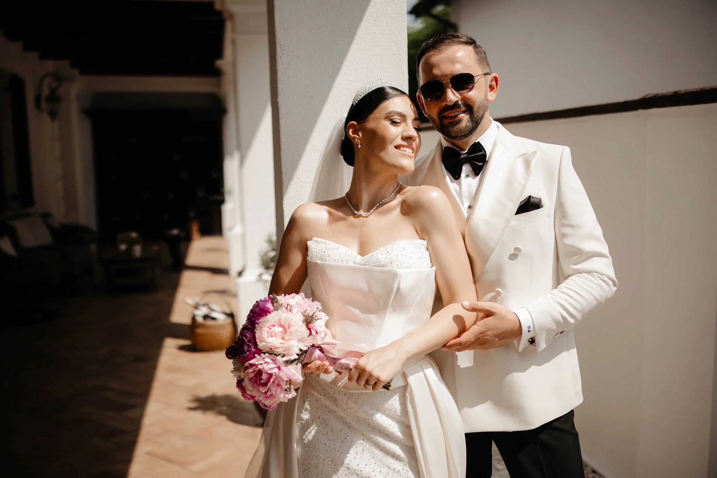 A bride and groom are embracing outdoors, dressed in wedding attire. The bride holds a bouquet of pink and white flowers. The groom is wearing sunglasses and a white tuxedo with a black bowtie. They are smiling and appear happy.