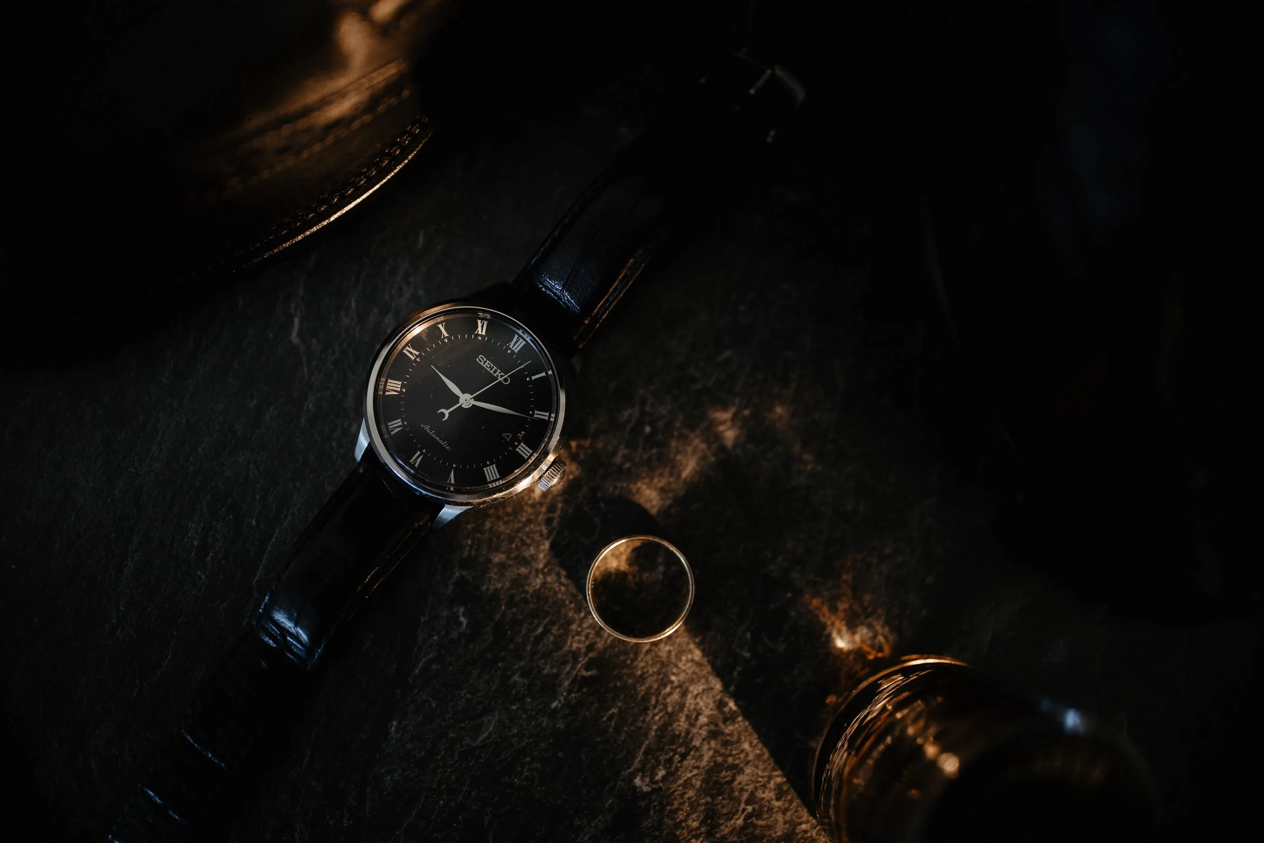 A black leather strap wristwatch with a black dial and Roman numerals, placed on a dark surface. Nearby is a gold ring and a glass of dark liquid