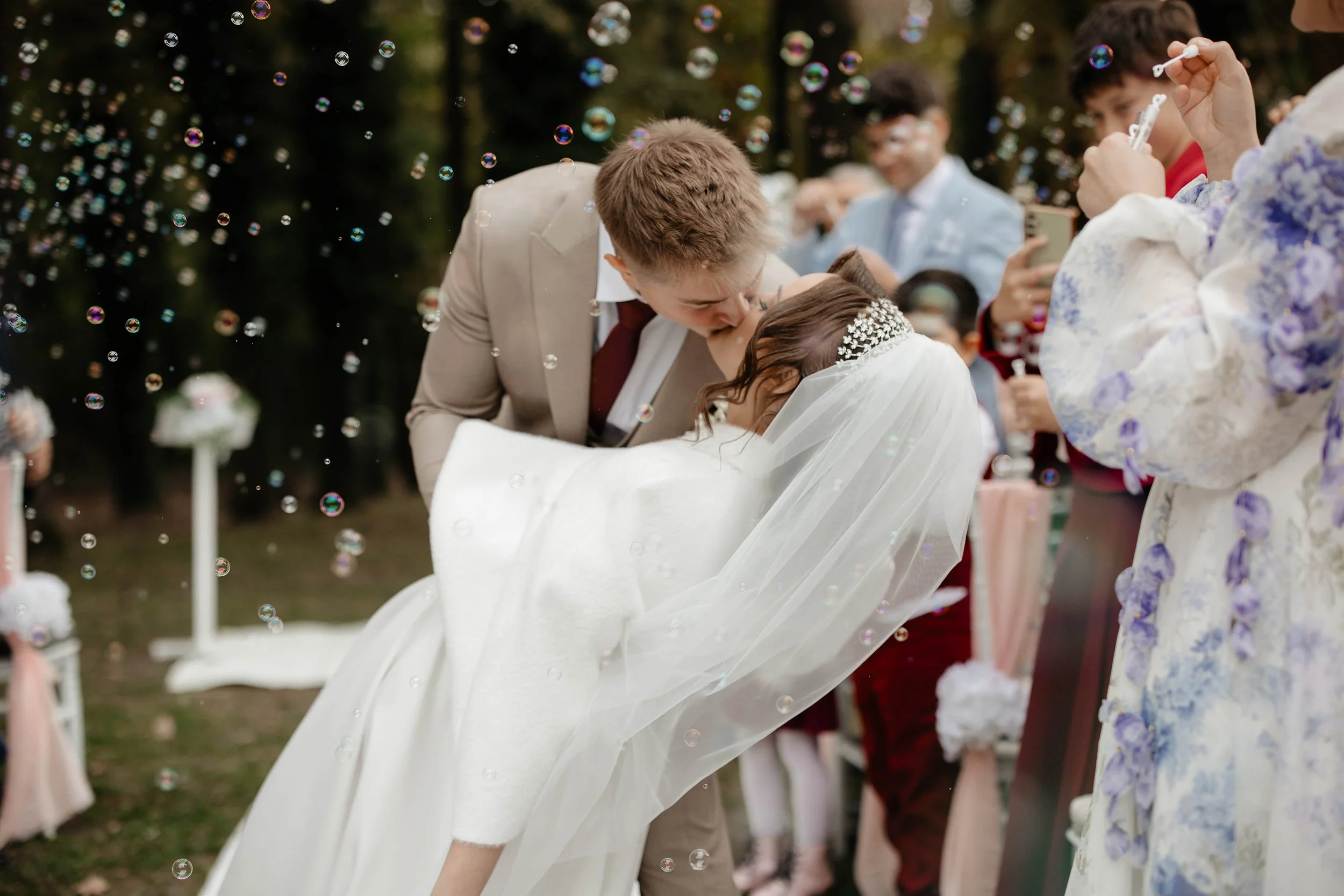 A bride and groom kiss during their outdoor wedding, surrounded by guests and bubbles floating in the air.